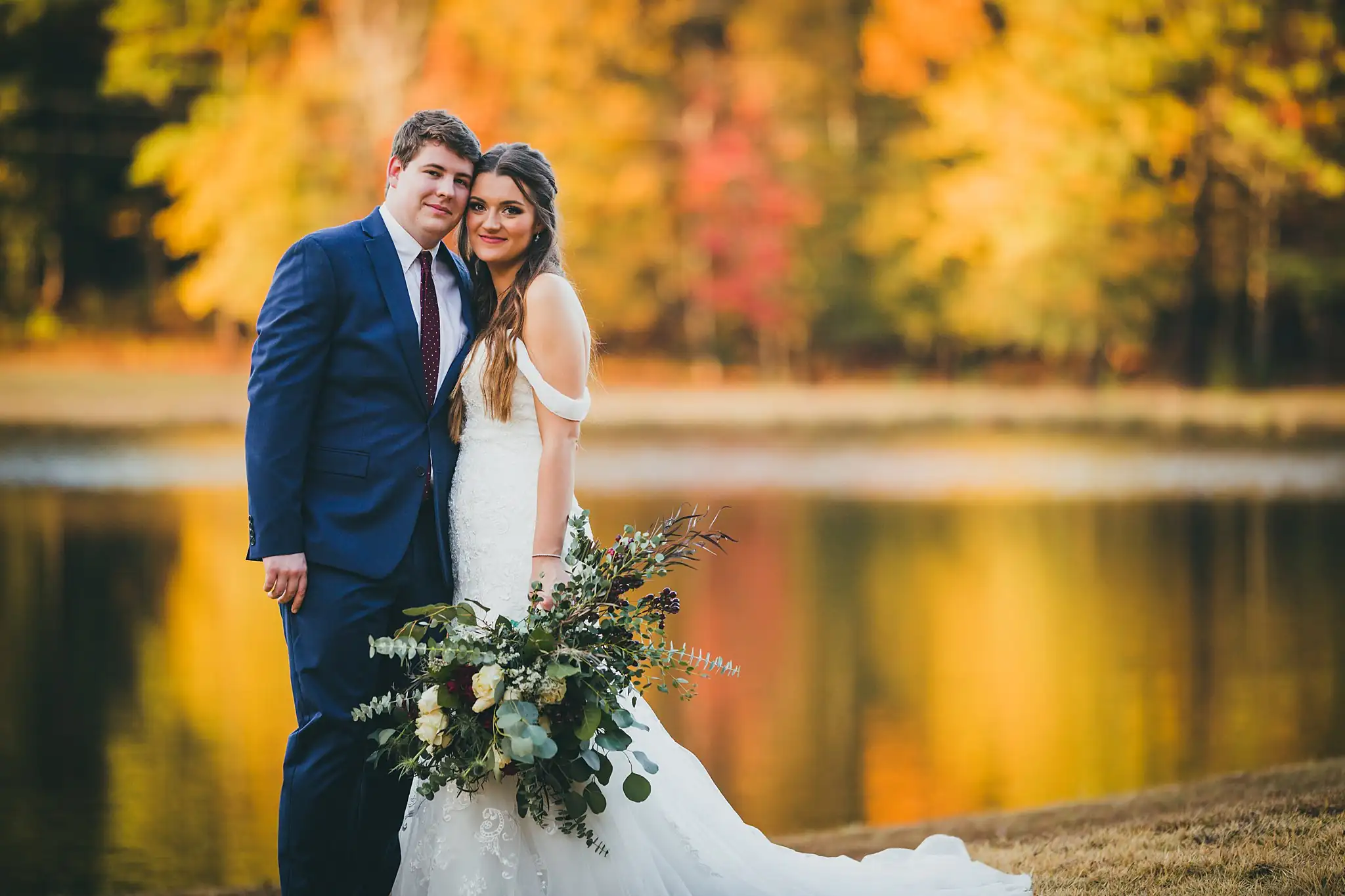 Barn at Harper Pines Wedding Thomaston, GA Atlanta Wedding Photographers Wedding Formals: the couple by the lake with autumn color behind them, at their wedding at the Barn at Harper Pines in Thomaston, GA