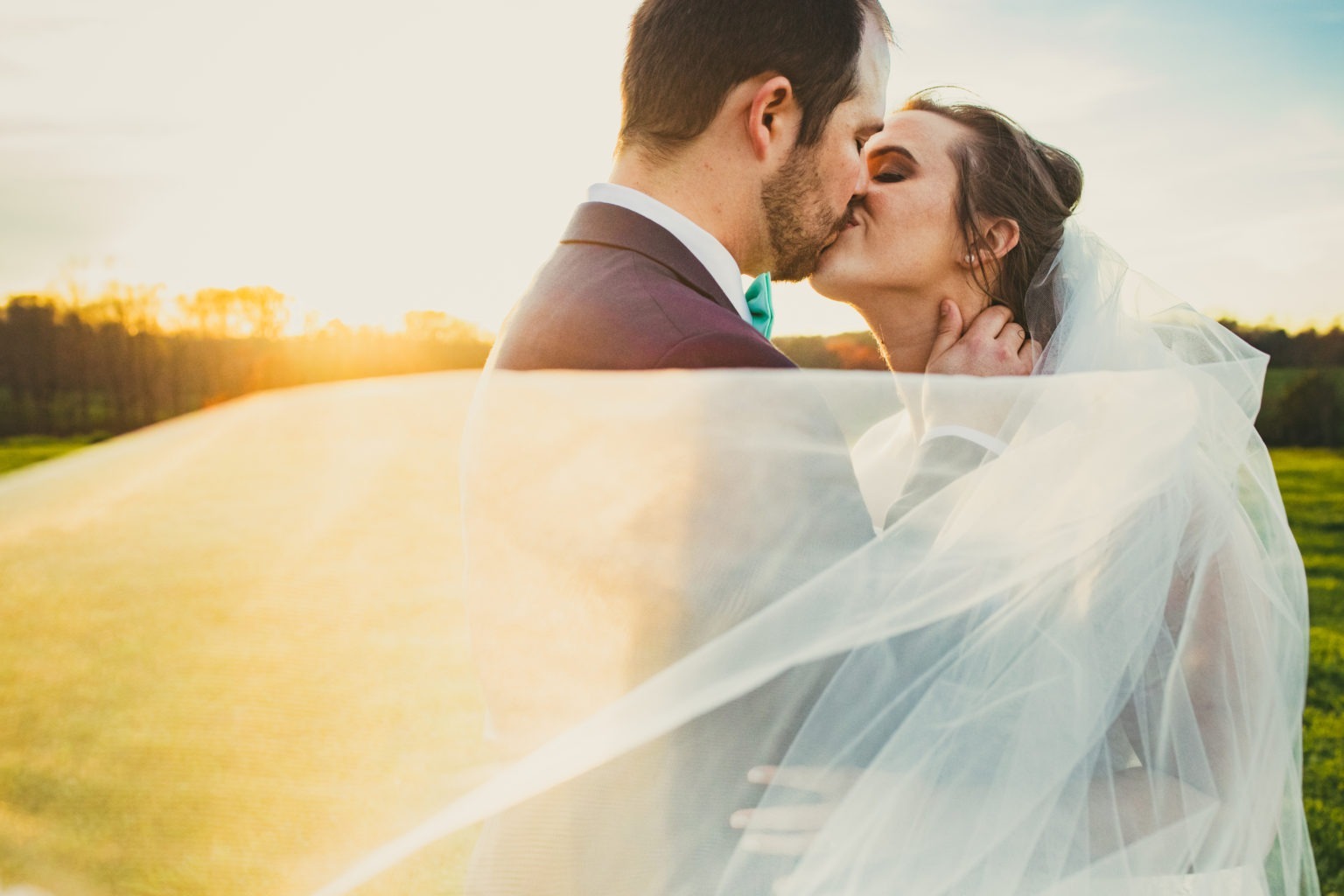 Bride and groom kissing under a billowing veil at sunset on their wedding day