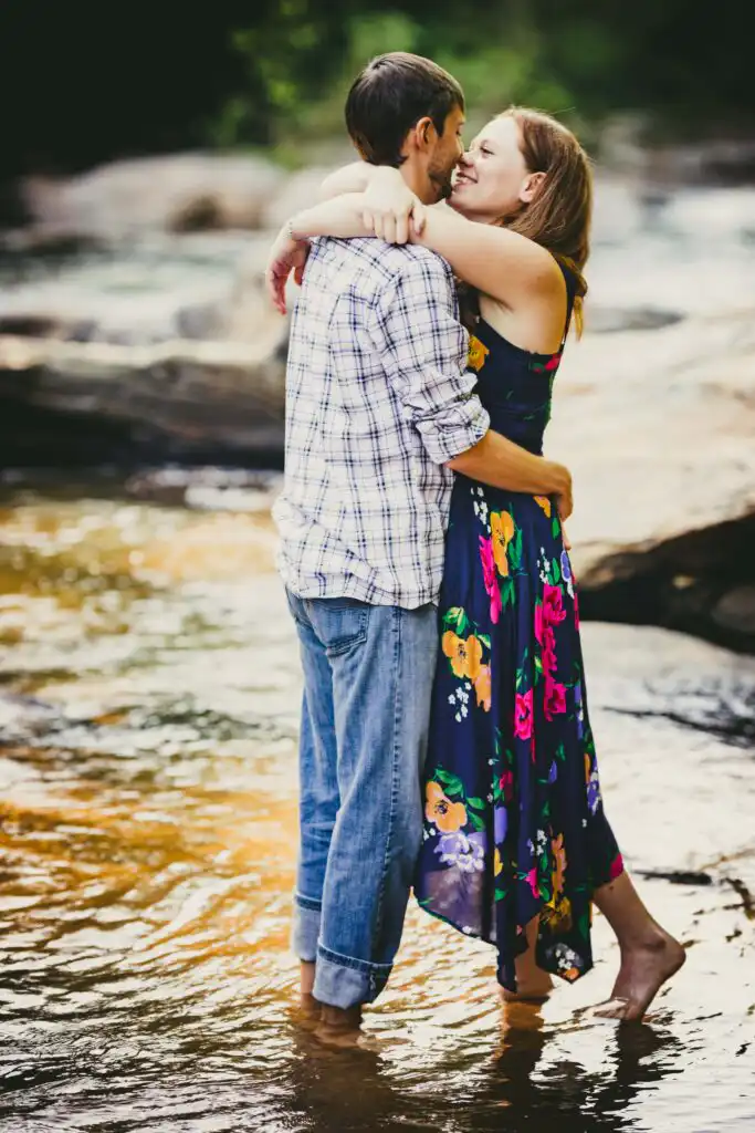 Engagement Portraits: a couple barefoot in a shallow river, her arms around his neck, during their engagement session at the Sope Creek mill ruins in Marietta, GA