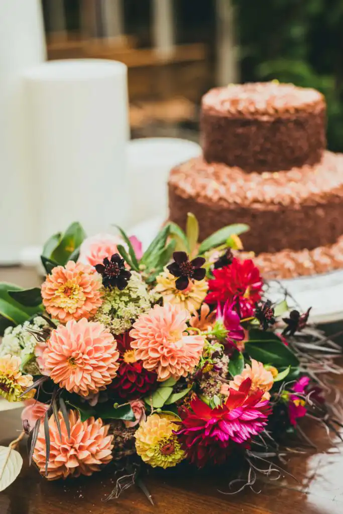 Wedding Details: a coral and magenta dahlia bouquet by Moss + Clay beside the tiered cake, at a wedding at Wahoo! Grill in Decatur, GA