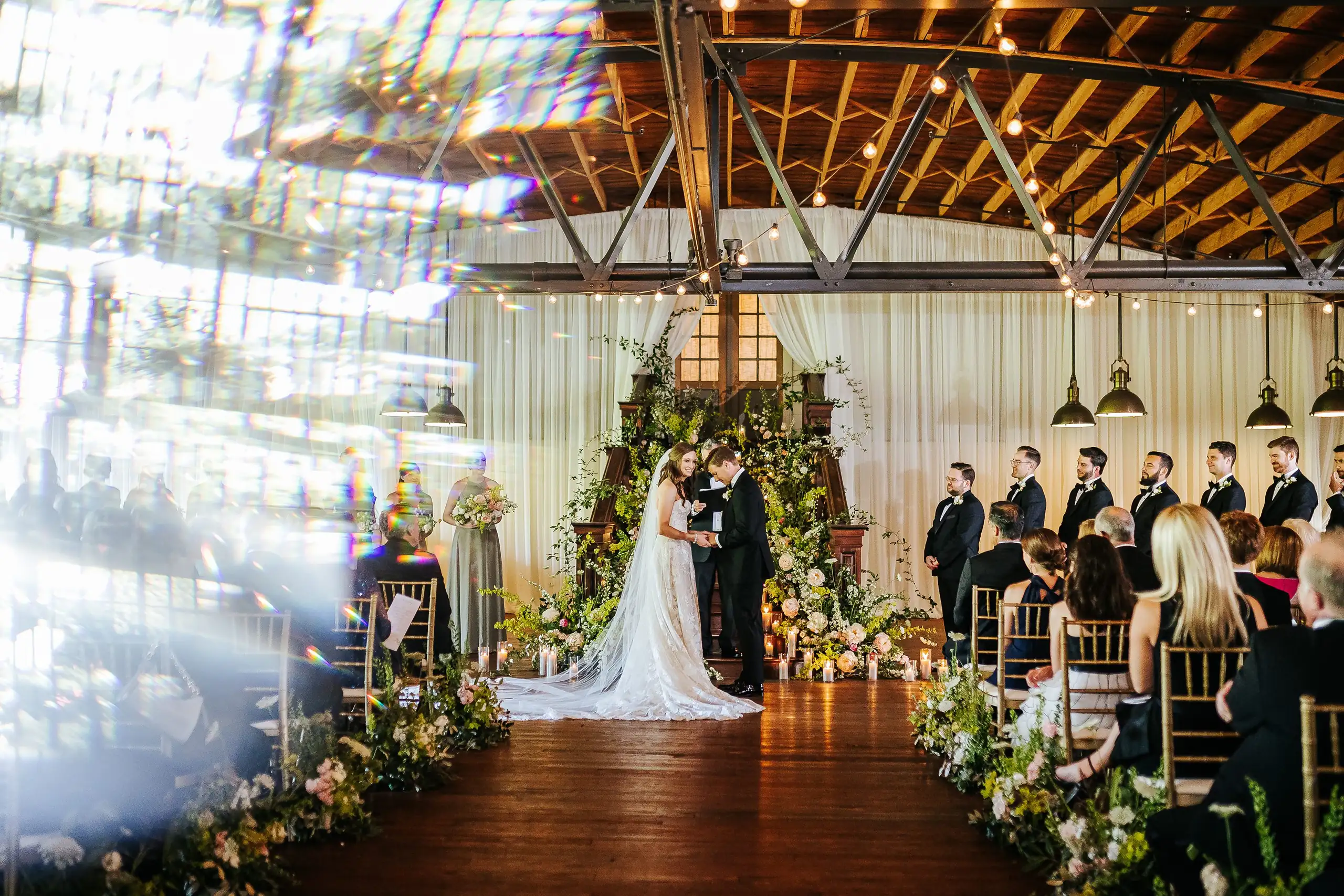 Wedding Ceremony: the ceremony beneath the timber trusses and floral arch, at their wedding at Summerour Studio in Atlanta, GA