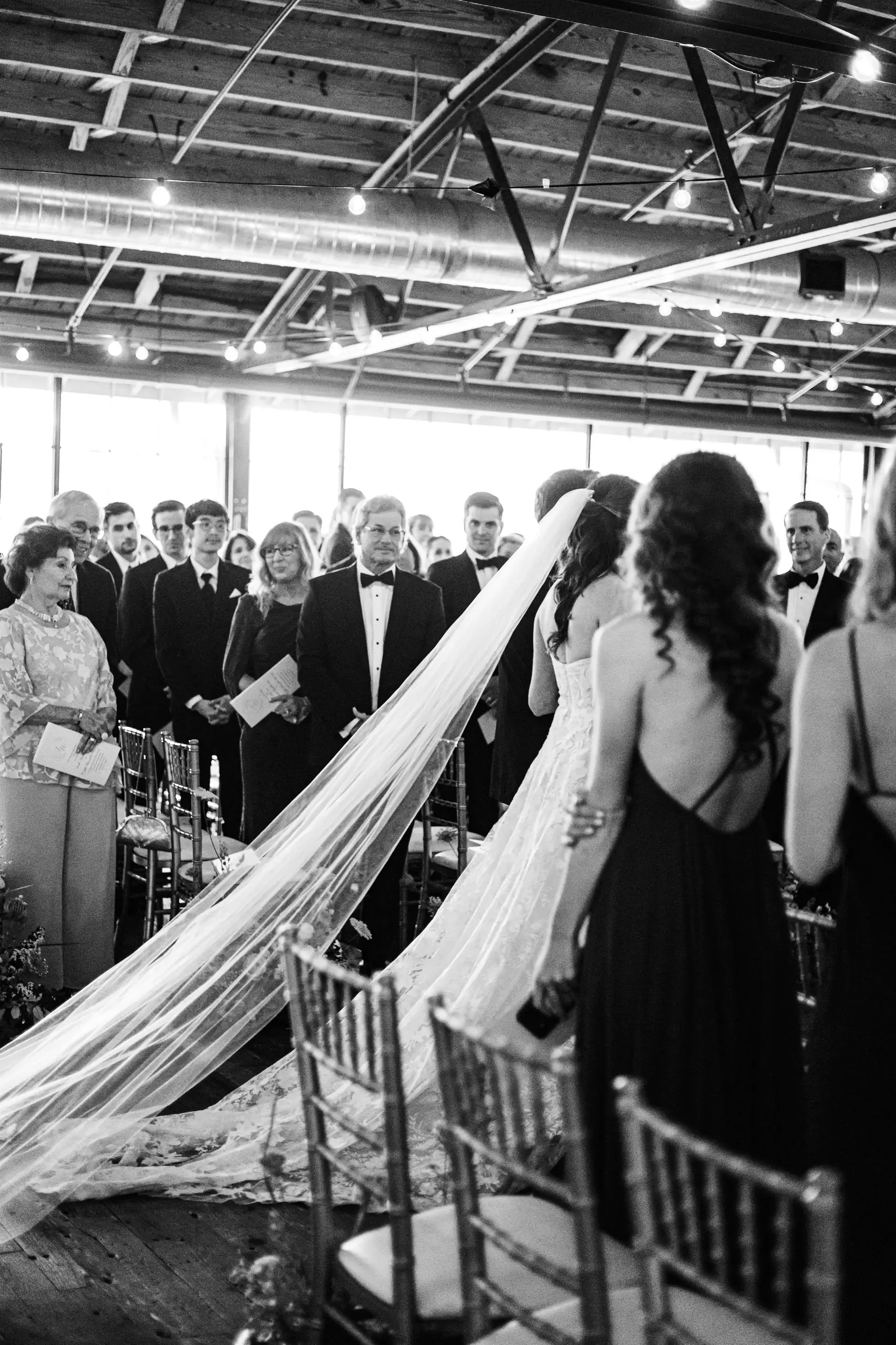Processional: the bride's cathedral veil trailing down the aisle, in black and white, at their wedding at Summerour Studio in Atlanta, GA