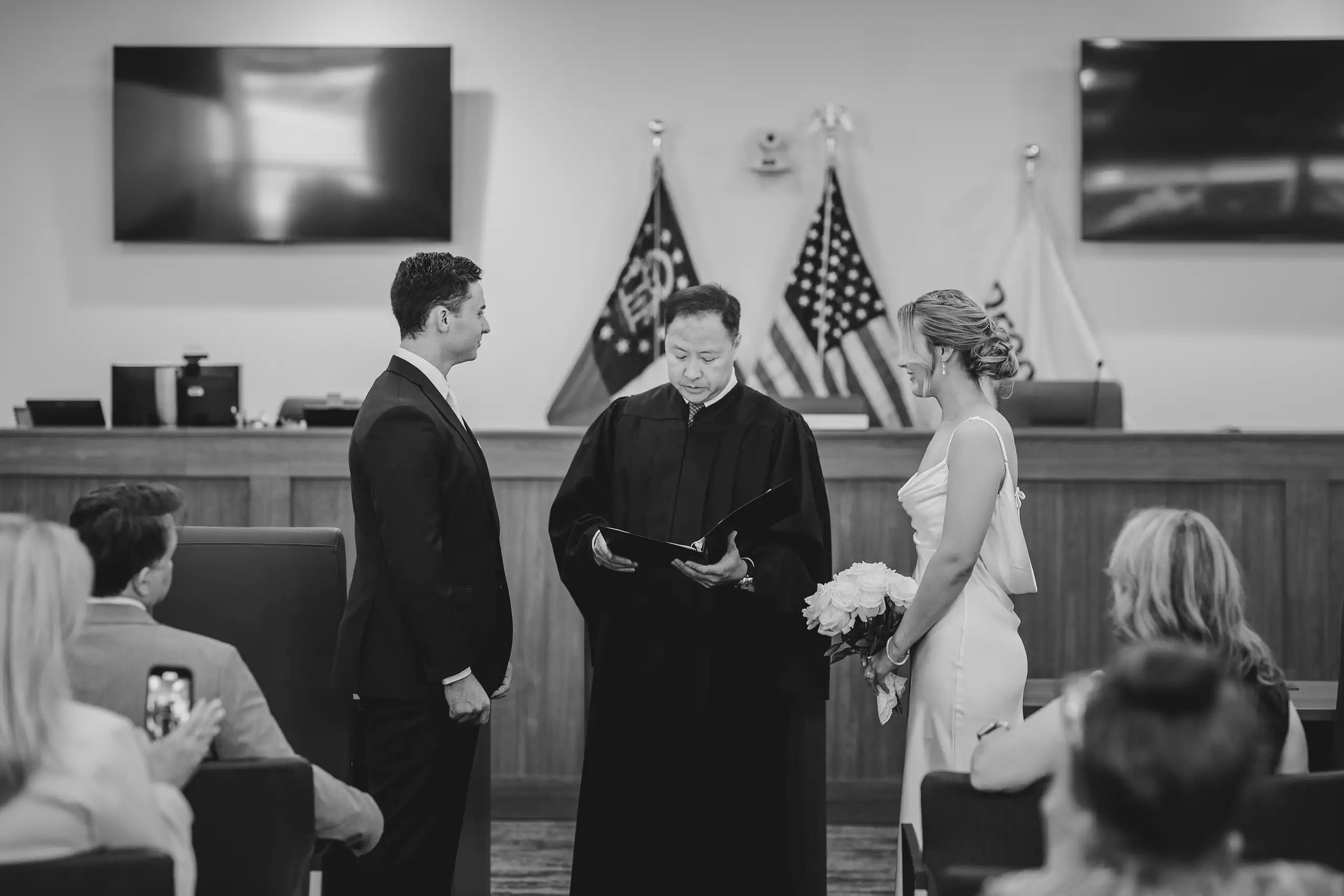 Wedding Ceremony: the couple before the judge between the flags, in black and white, at their courthouse wedding in Brookhaven, GA