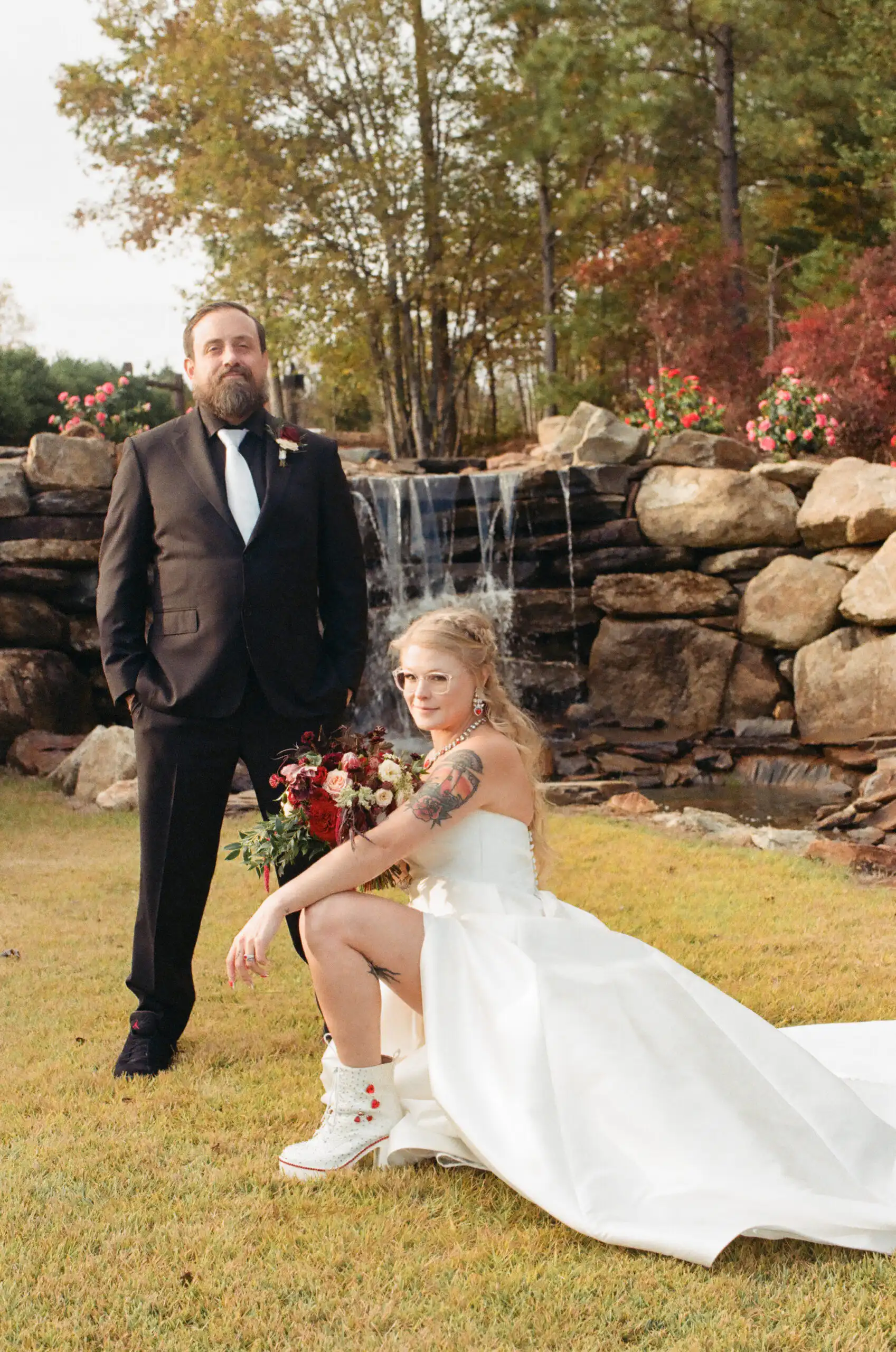 Bride seated on the lawn with her bouquet and the groom beside a waterfall at their wedding, shot on 35mm film at The Venue at Sharp Mountain in North Georgia