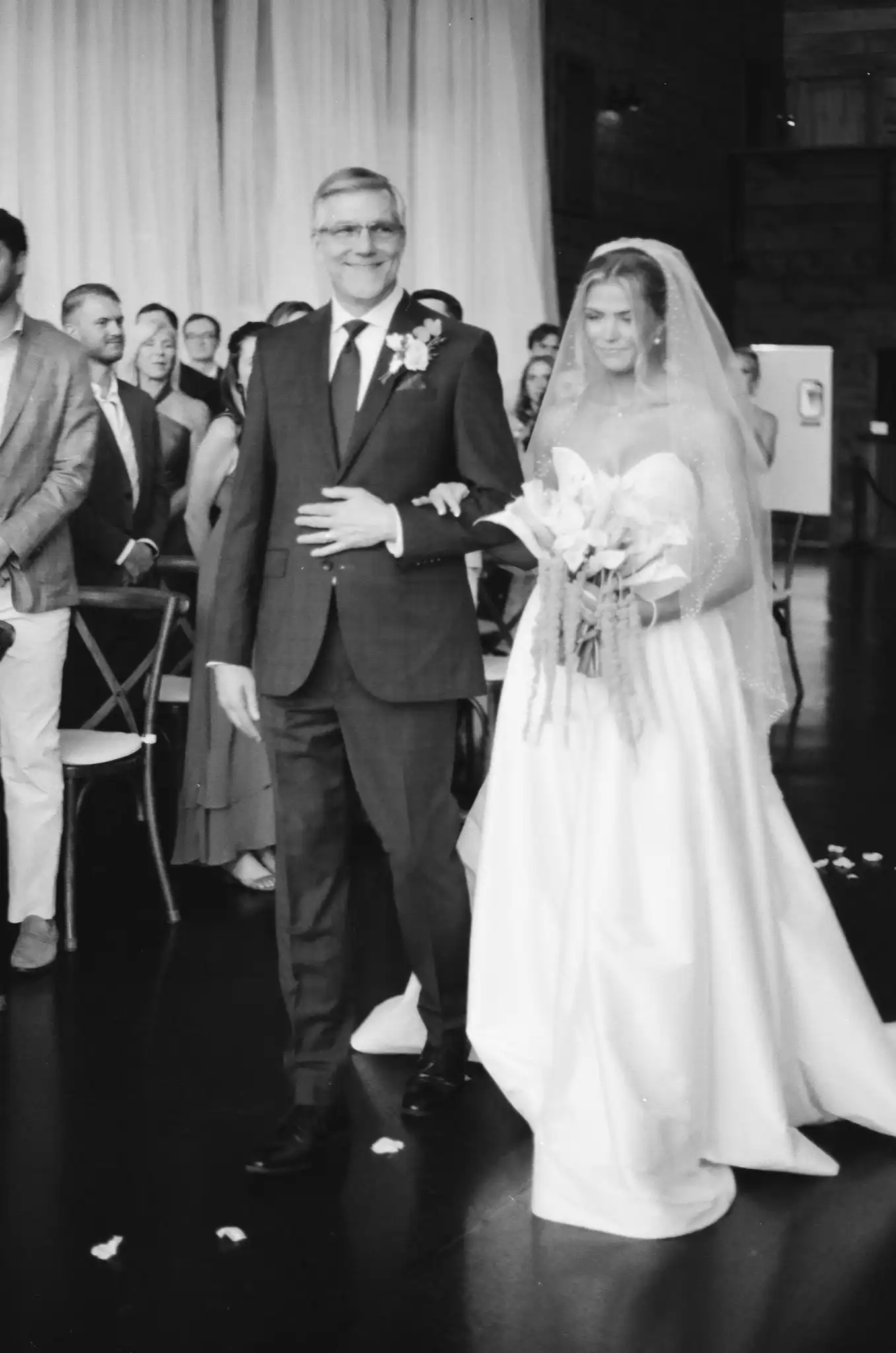 Bride walking down the aisle with her father past the guests, in black and white on 35mm film at The Venue at Sharp Mountain in Jasper, Georgia