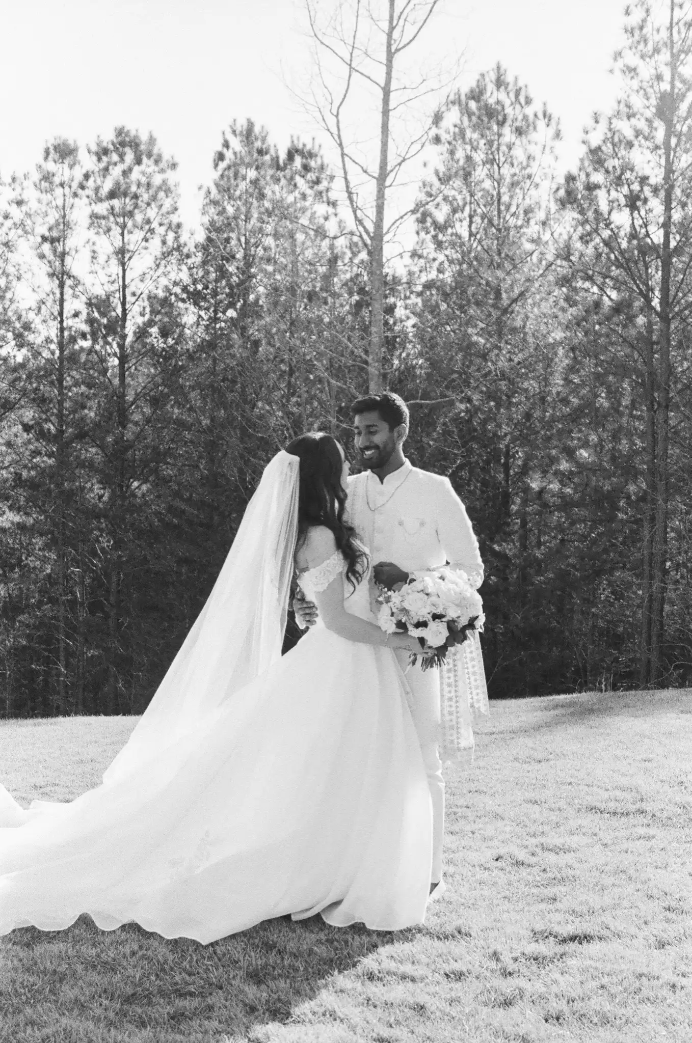 Bride in a cathedral veil and groom in a sherwani among the pines, in black and white on 35mm film at The Venue at Sharp Mountain in Jasper, Georgia