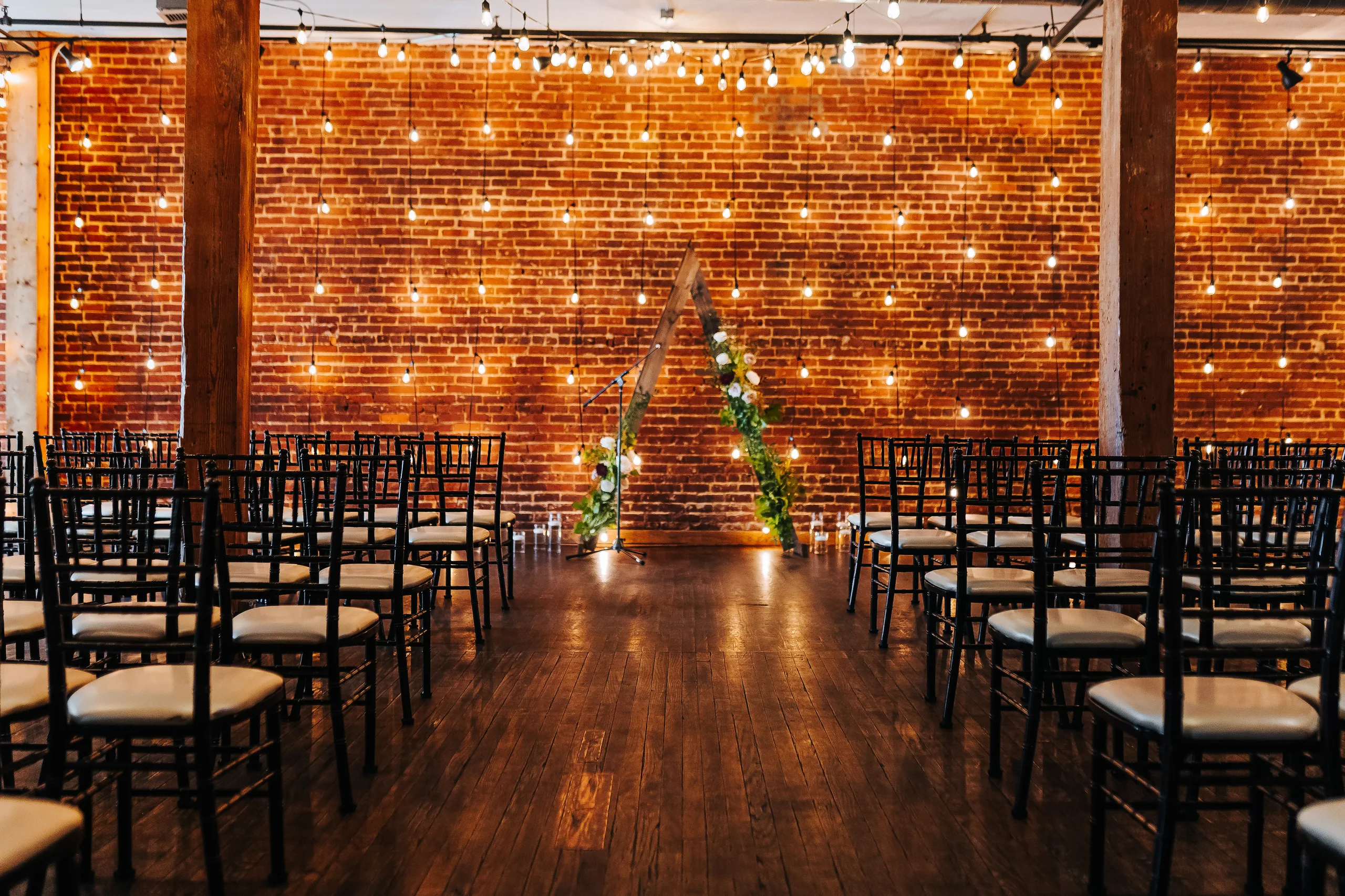 Wedding Ceremony: the brick wall hung with string lights and a wooden arch, chairs set on the hardwood floor, at a wedding at Terminus 330 in Atlanta, GA