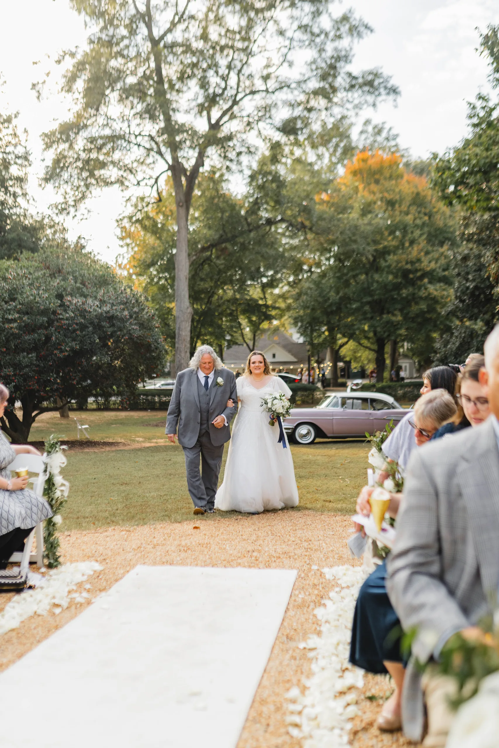 Wedding Ceremony: the bride coming down the aisle with her dad, the pink Chevrolet behind her, at their wedding at Naylor Hall in Roswell, GA