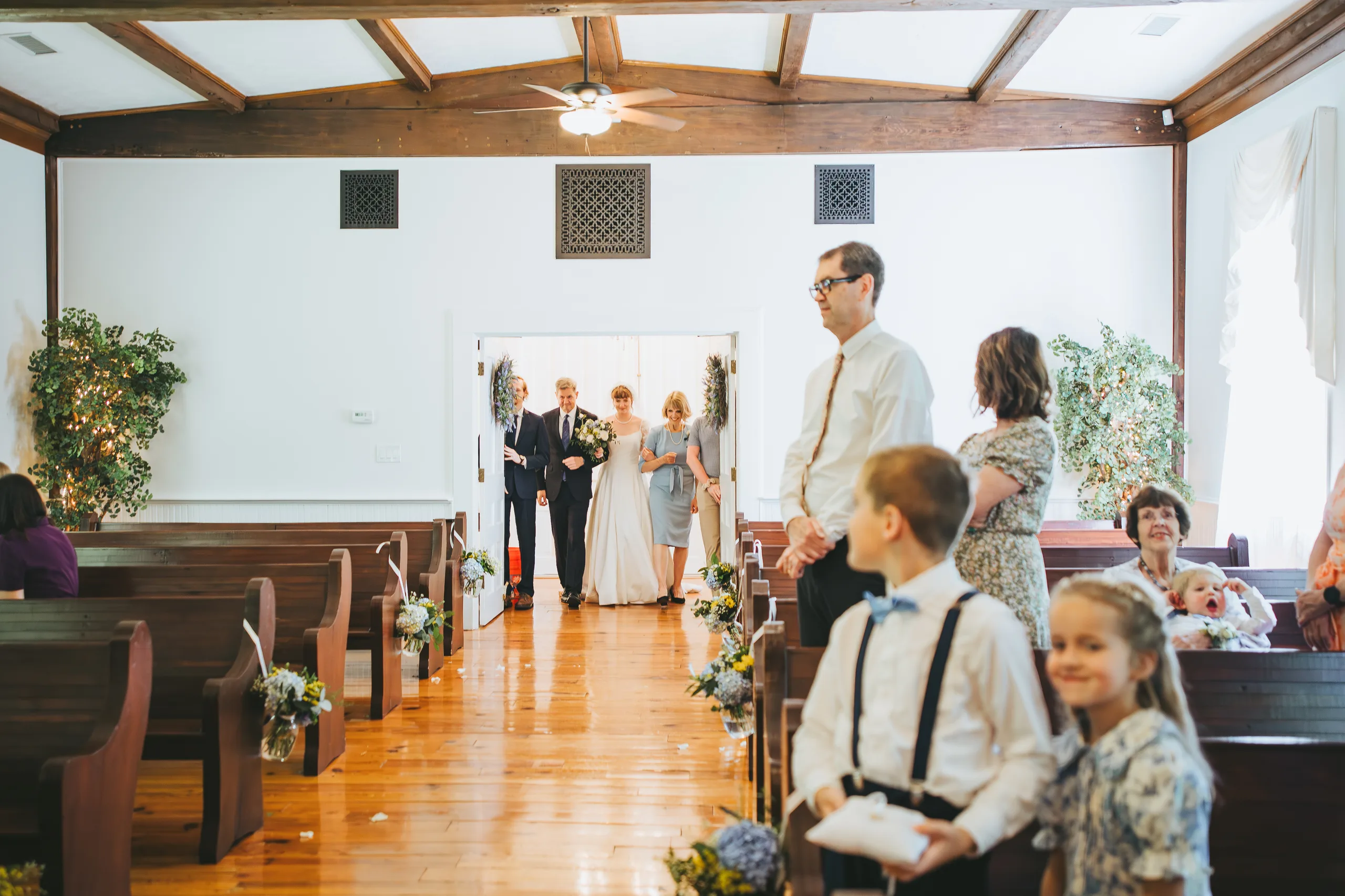 Wedding Ceremony: the bride walking in with guests turning to look, at their wedding at the Chapel at Simpsonwood Park in Peachtree Corners, GA