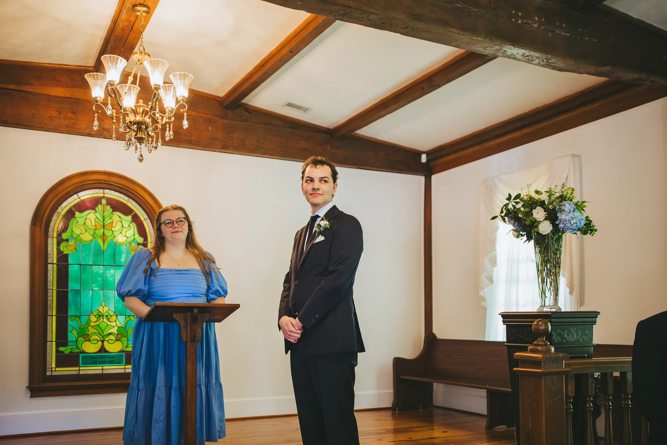 Wedding Ceremony: the groom waiting at the front by the stained glass window, at their wedding at the Chapel at Simpsonwood Park in Peachtree Corners, GA