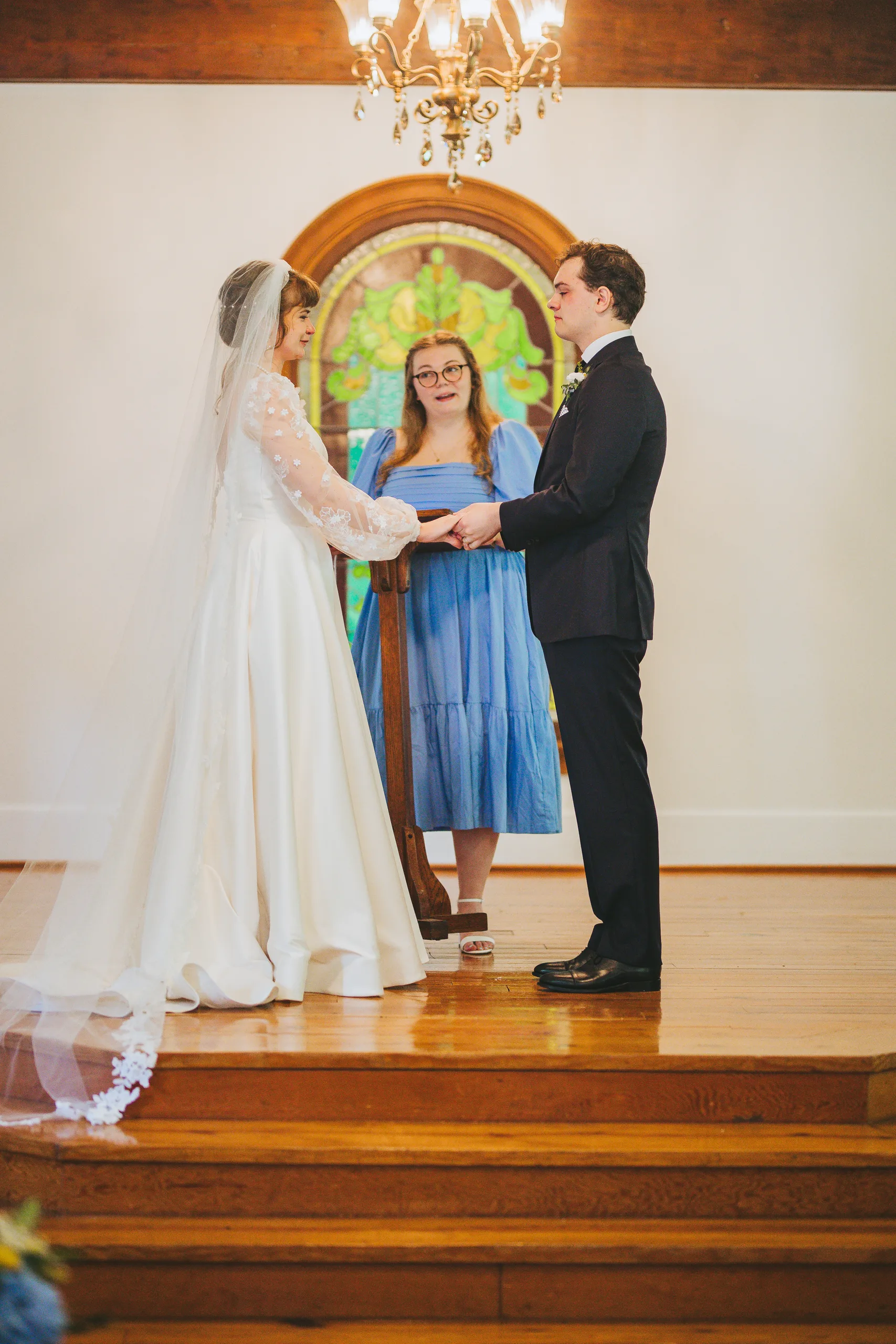Wedding Ceremony: exchanging vows in front of the stained glass window, at their wedding at the Chapel at Simpsonwood Park in Peachtree Corners, GA