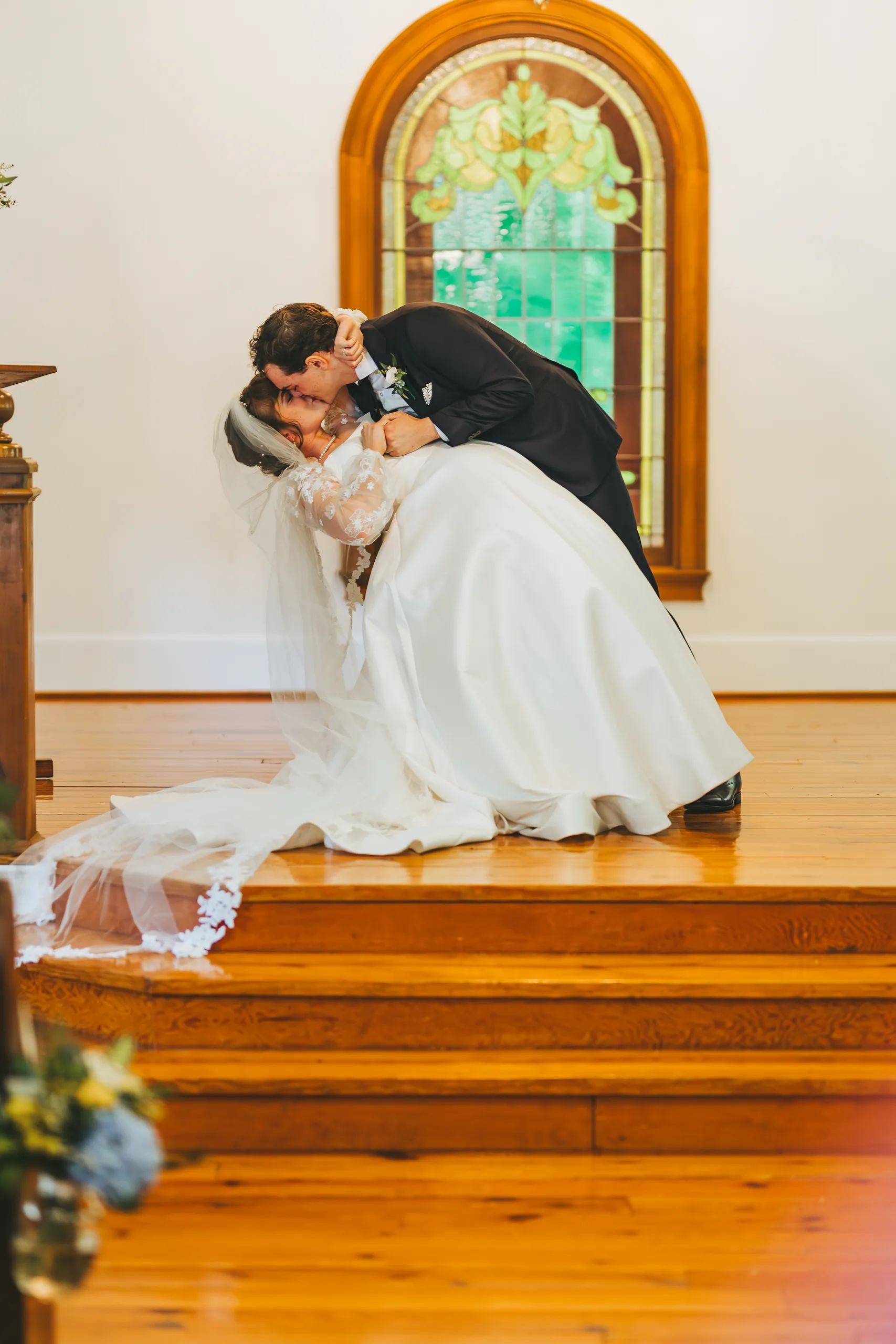 Wedding Ceremony: a dip kiss in front of the stained glass window, at their wedding at the Chapel at Simpsonwood Park in Peachtree Corners, GA