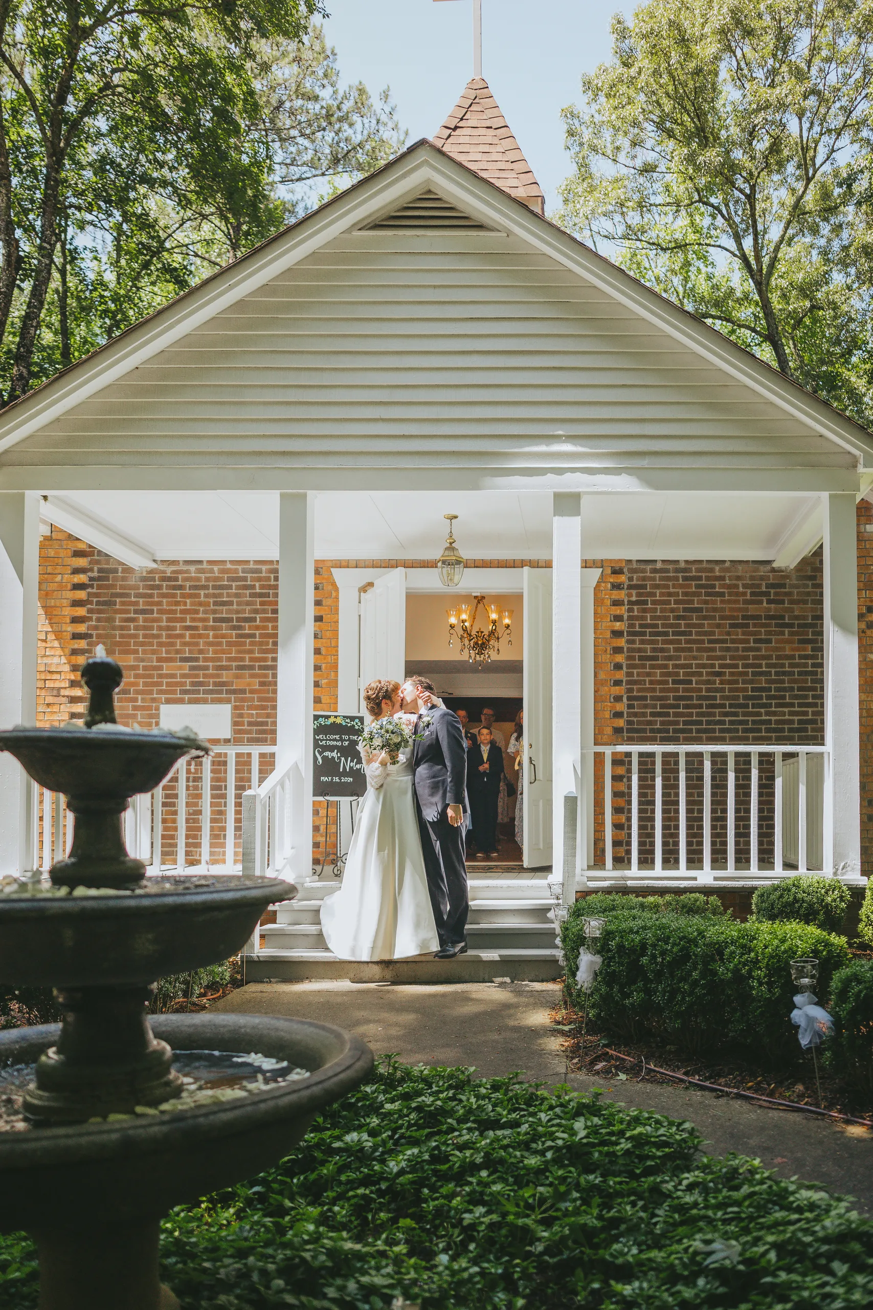 Wedding Ceremony: a kiss on the chapel porch behind the fountain, at their wedding at the Chapel at Simpsonwood Park in Peachtree Corners, GA
