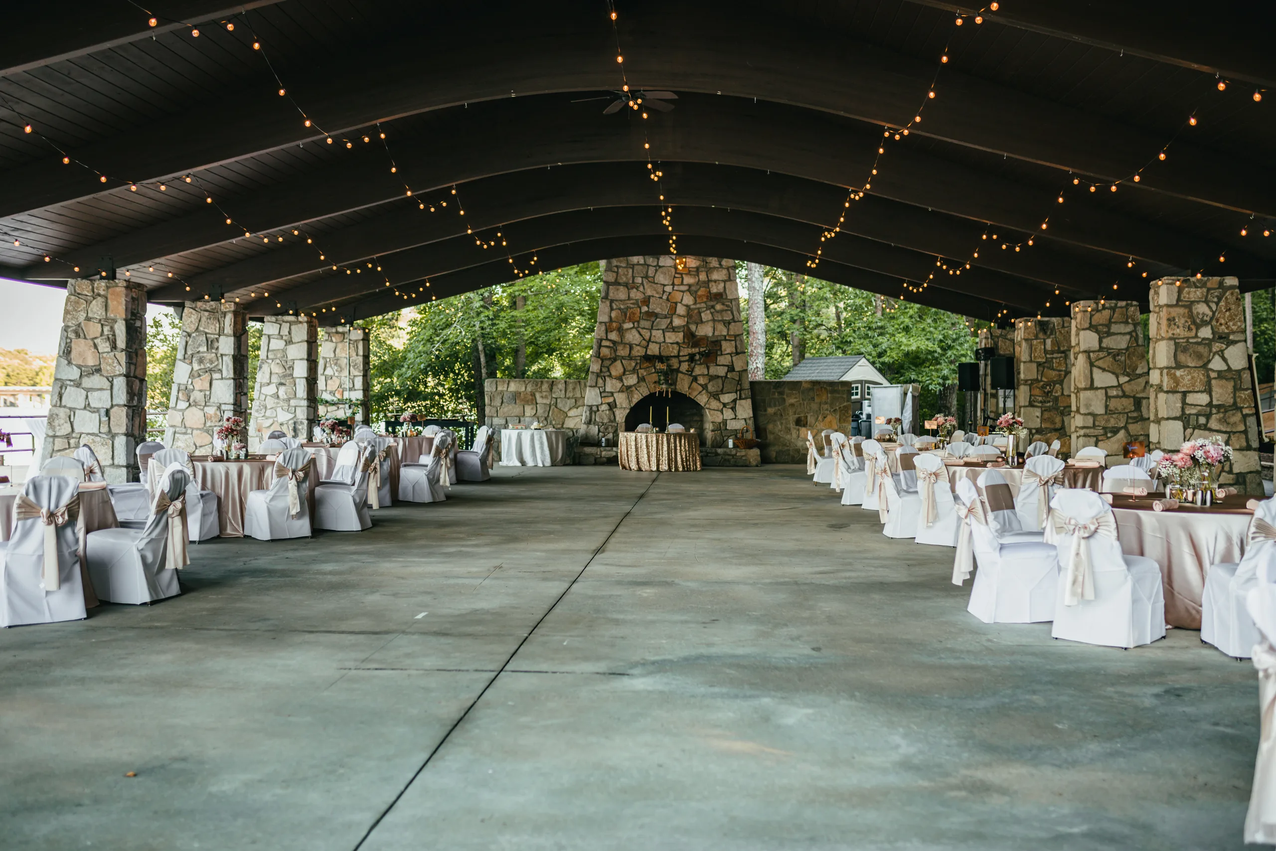 Reception Details: the pavilion's big stone fireplace and the tables under string lights, at a wedding at Atlanta Evergreen Lakeside Resort in Stone Mountain, GA