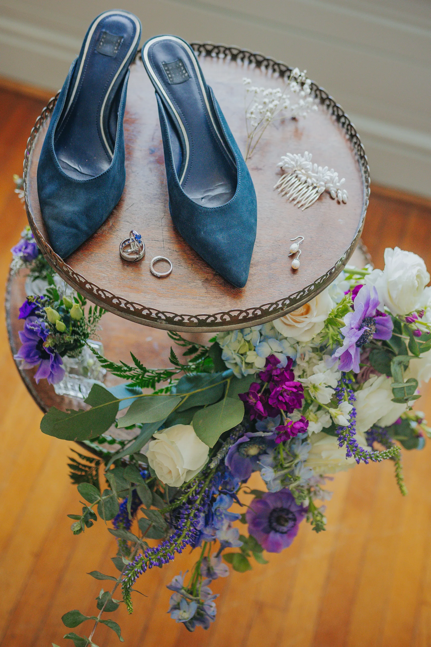 Wedding Details: her blue suede heels, rings and hairpieces on a tiered stand with purple and white flowers, at a wedding at The Wildflower 301 in Greensboro, GA