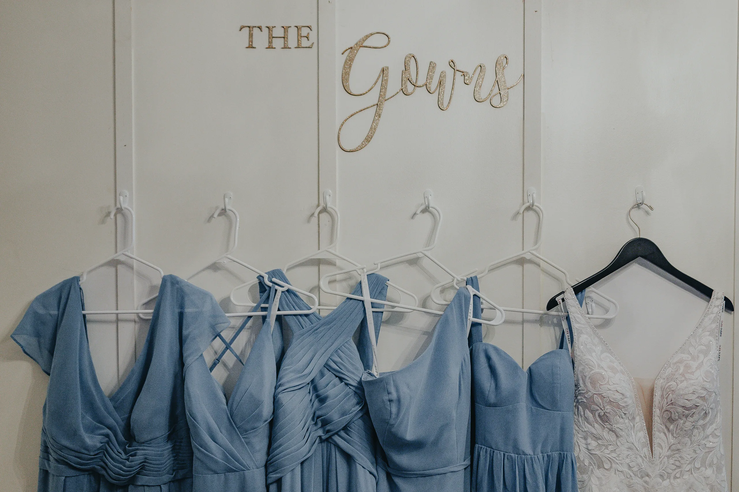 Wedding Details: the dusty blue bridesmaids' dresses and her gown hanging under a The Gowns sign, at a wedding at The Wildflower 301 in Greensboro, GA