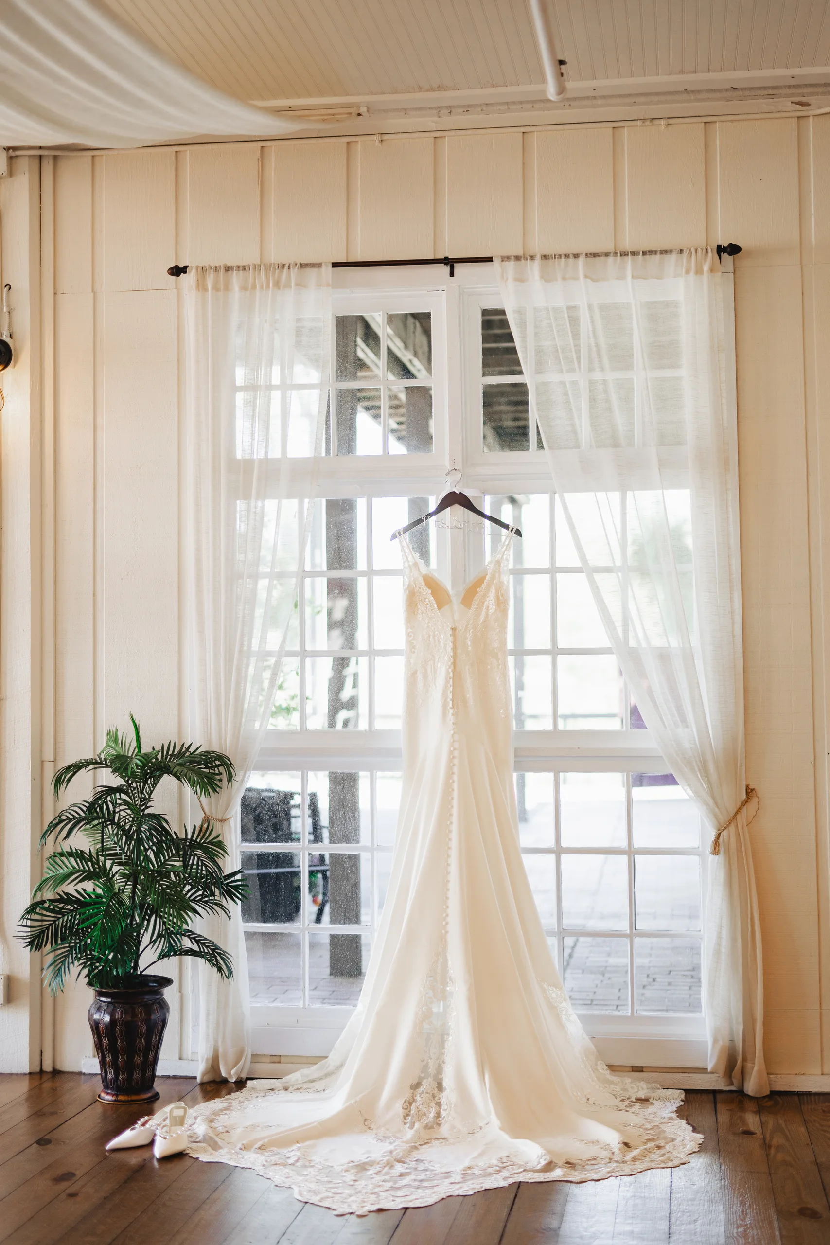 Wedding Details: her gown in the bridal suite window beside a potted palm before their wedding at Ivy Hall at Roswell Mill in Roswell, GA