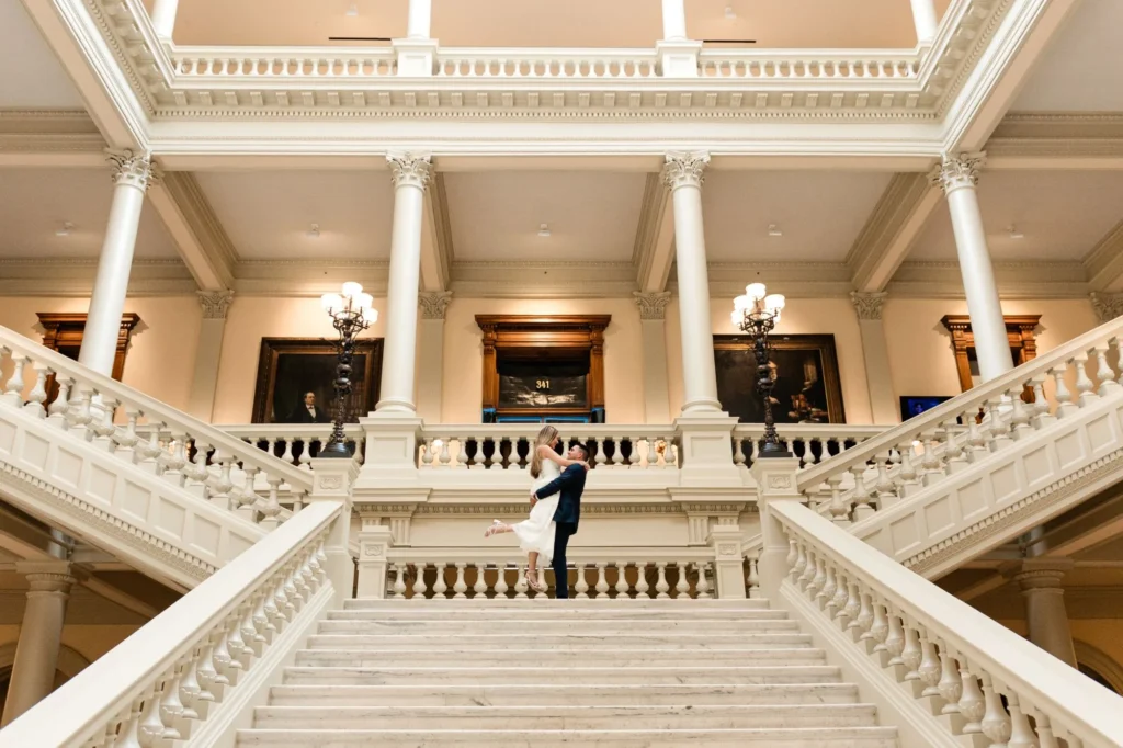 Grand Staircase: he lifts her at the top of the marble staircase, her heel kicked up, columns and portraits above, at their engagement session at the Georgia State Capitol in Atlanta, GA