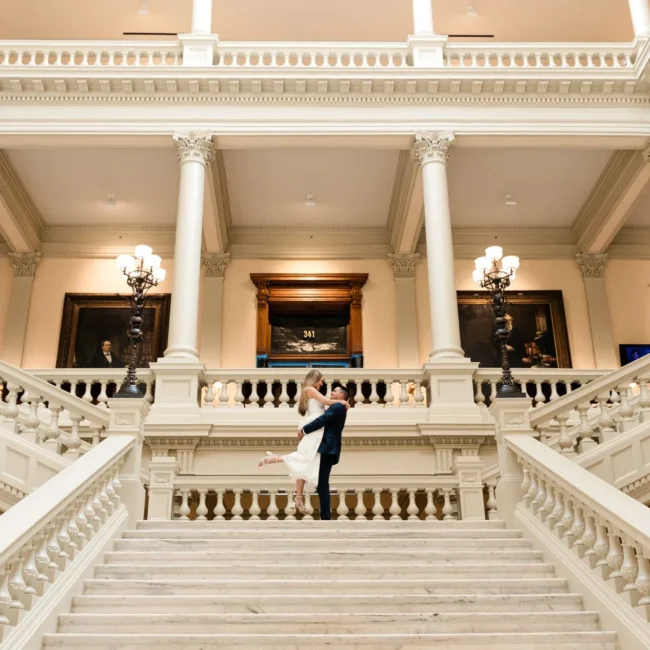 Grand Staircase: he lifts her at the top of the marble staircase, her heel kicked up, columns and portraits above, at their engagement session at the Georgia State Capitol in Atlanta, GA