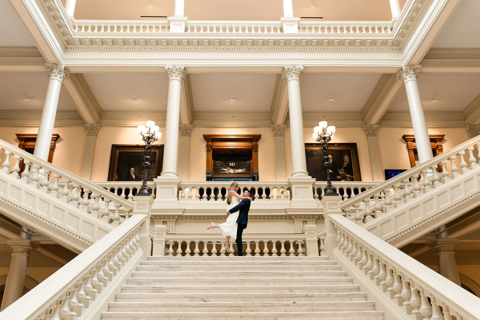 Grand Staircase: he lifts her at the top of the marble staircase, her heel kicked up, columns and portraits above, at their engagement session at the Georgia State Capitol in Atlanta, GA