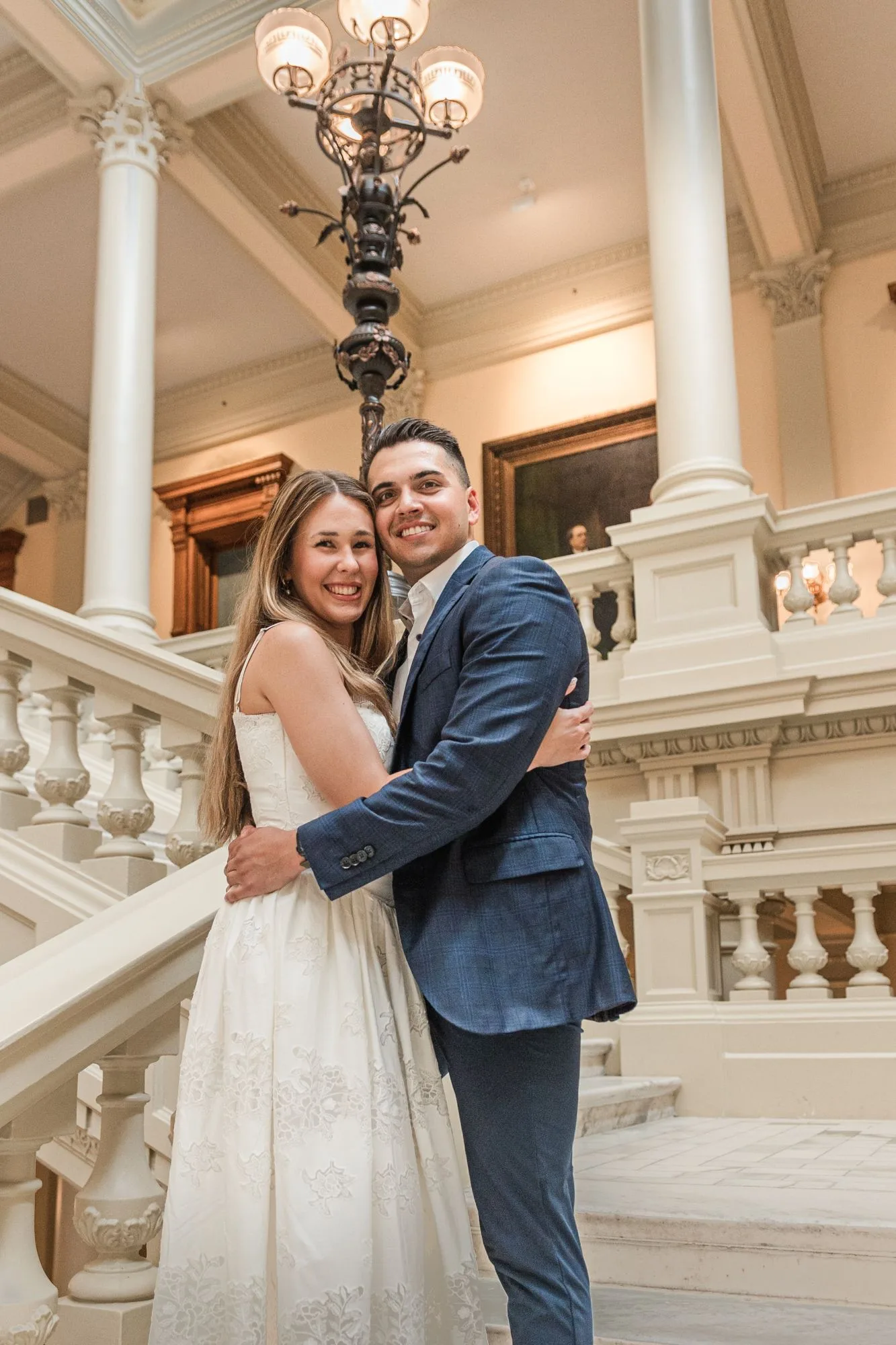 Grand Staircase: the two of them hugging and smiling on the stairs under a bronze lamp, at their engagement session at the Georgia State Capitol in Atlanta, GA