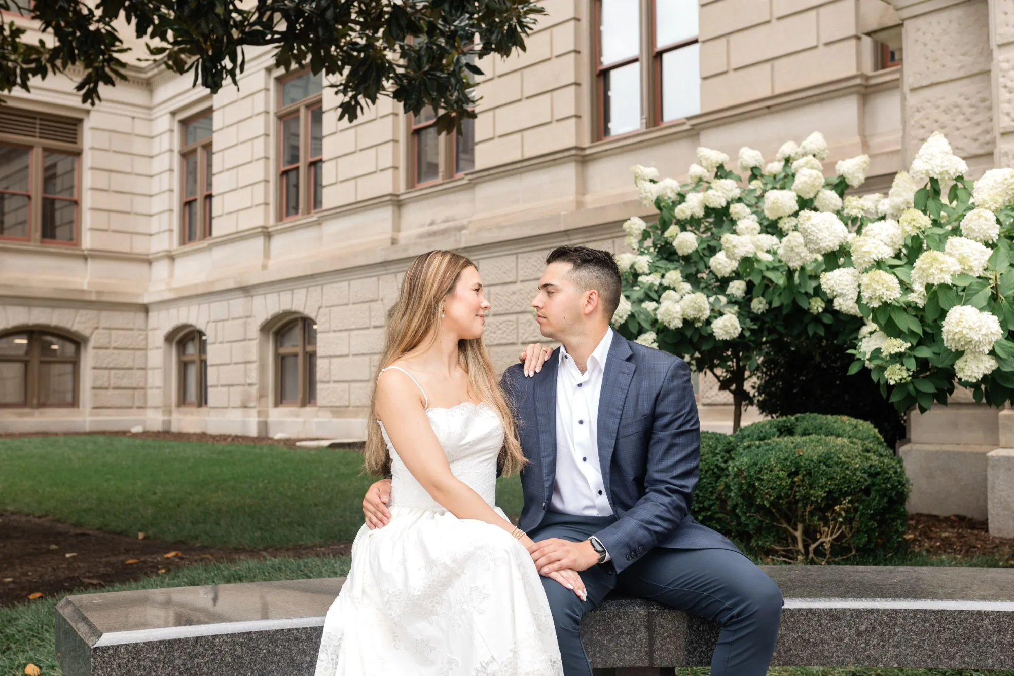Capitol Grounds: sitting on a granite bench in front of white hydrangeas, looking at each other, at their engagement session at the Georgia State Capitol in Atlanta, GA