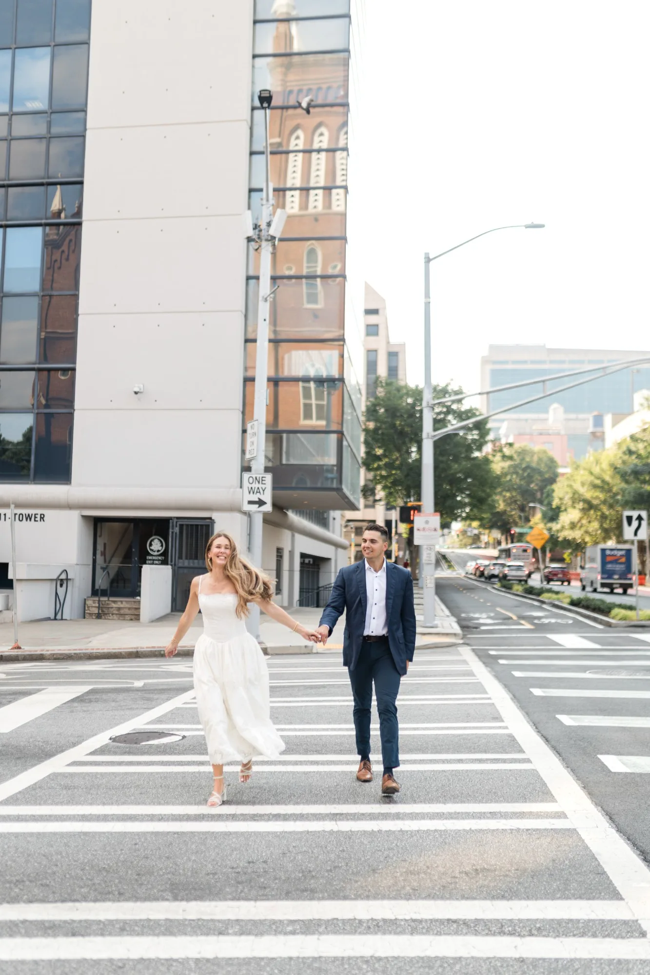 Downtown Atlanta: running across the crosswalk hand in hand, a church steeple reflected in the glass behind them, at their engagement session near the Georgia State Capitol in Atlanta, GA
