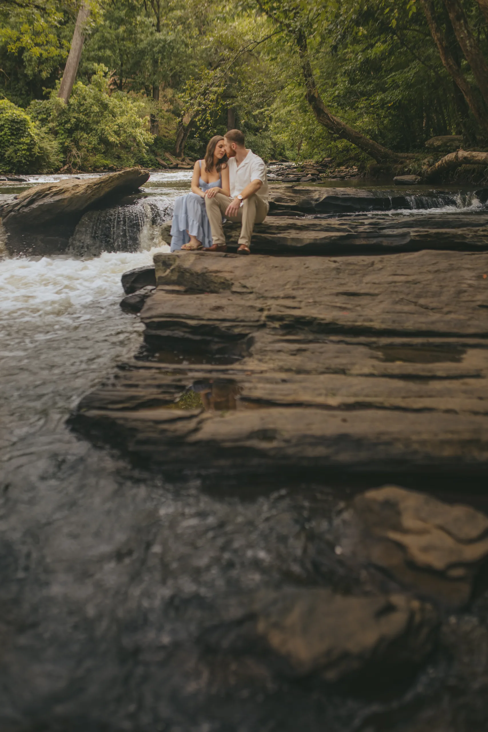 Engagement: sitting close on the rocks with the creek in front, at their engagement session at Roswell Mill in Roswell, GA