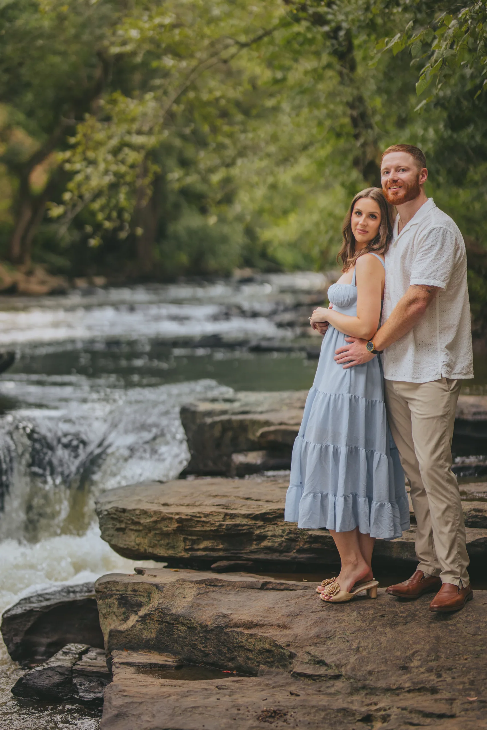 Engagement: standing on the rocks by the creek with the rapids beside them, at their engagement session at Roswell Mill in Roswell, GA
