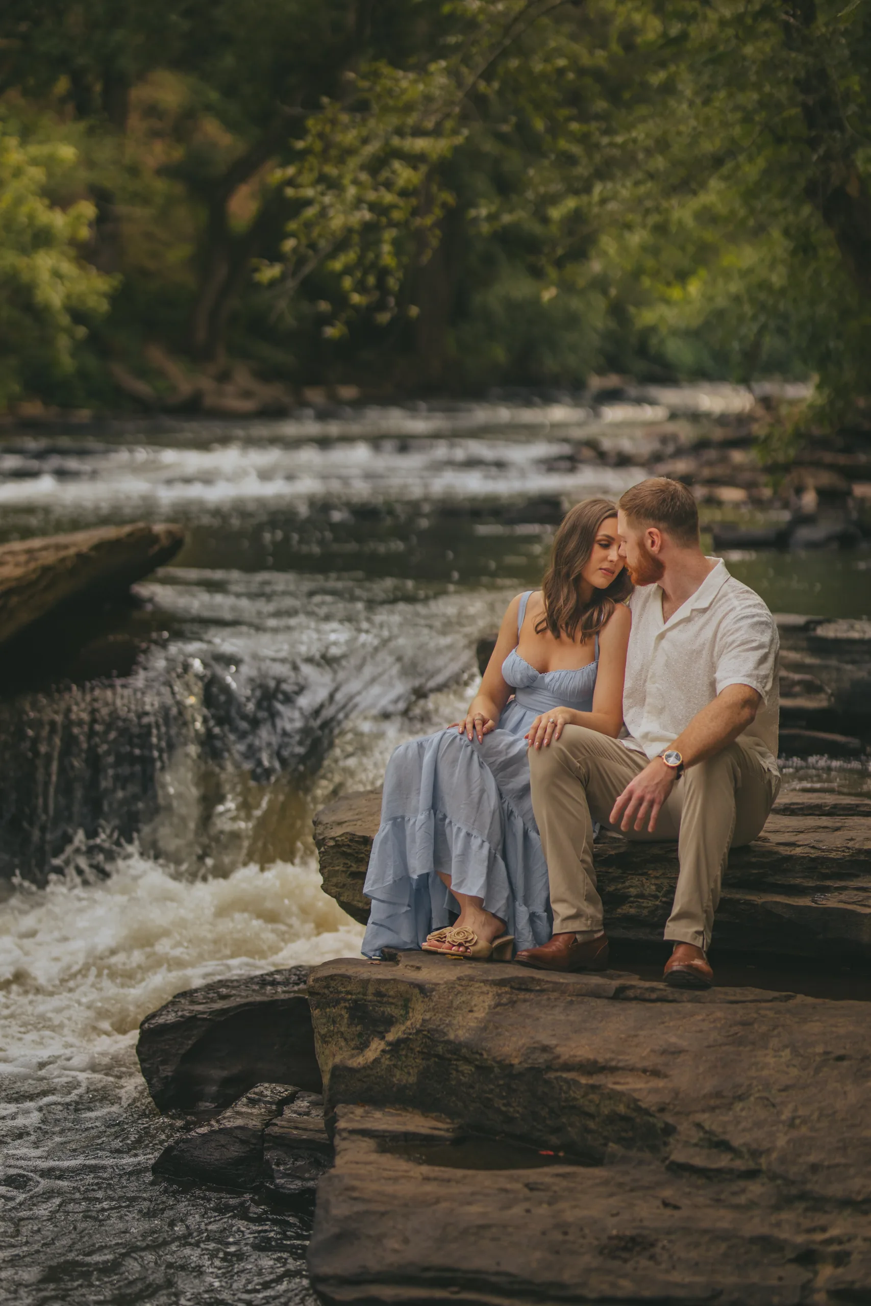 Engagement: sitting on the rocks by the water, at their engagement session at Roswell Mill in Roswell, GA