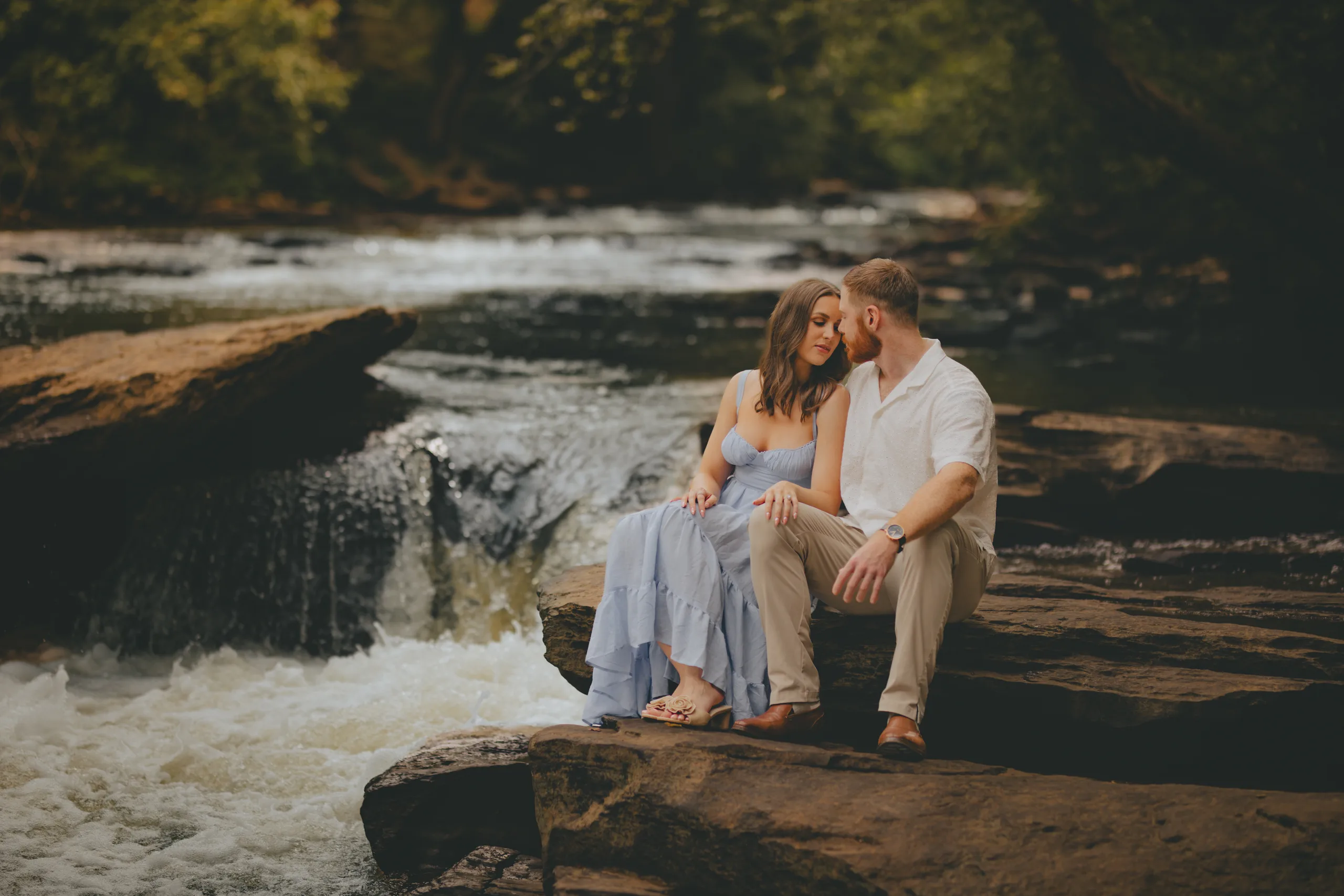 Engagement: sitting on the rocks beside a small waterfall, at their engagement session at Roswell Mill in Roswell, GA