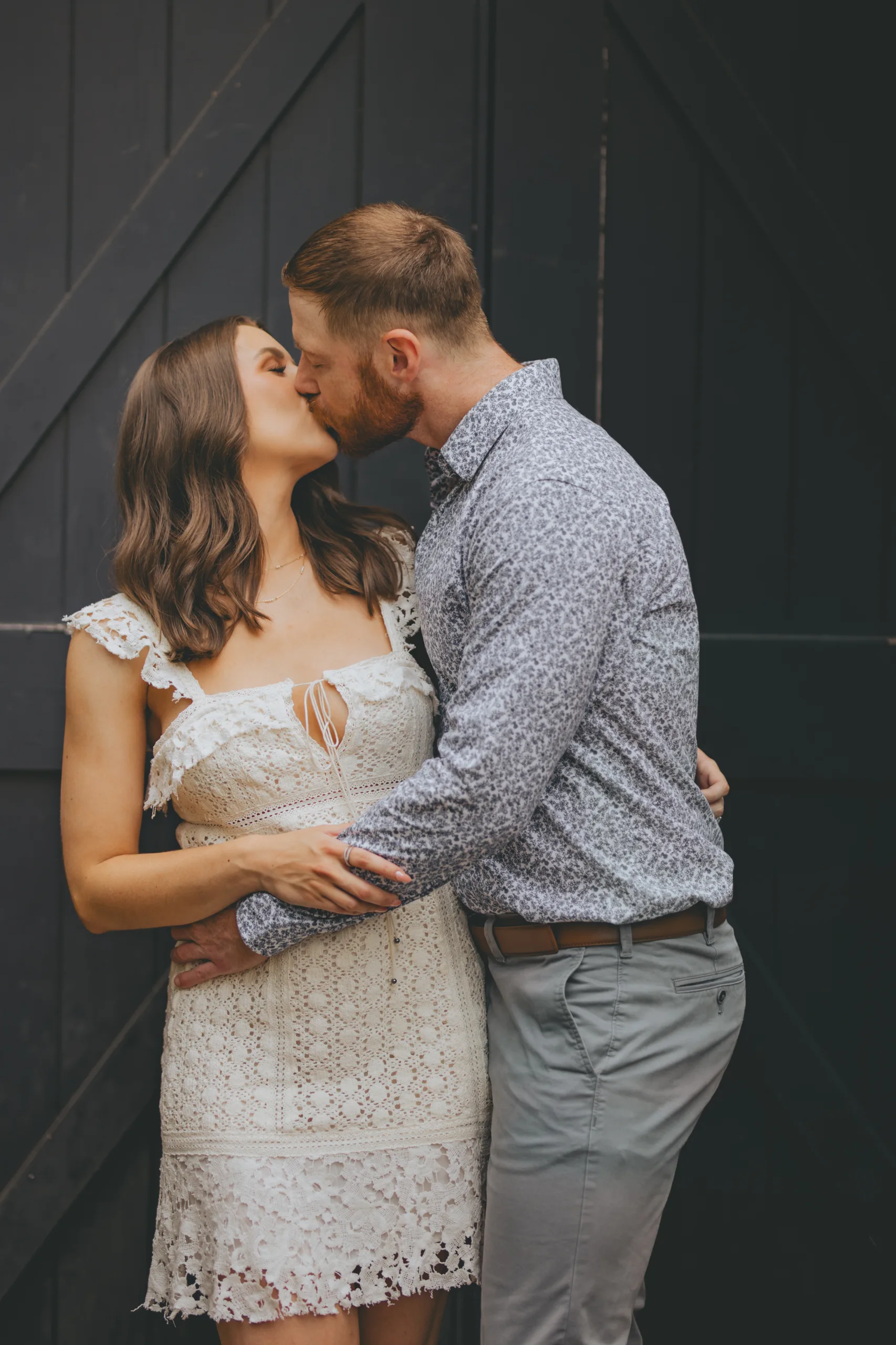 Engagement: a kiss in front of black barn doors, her in white lace, at their engagement session at Roswell Mill in Roswell, GA