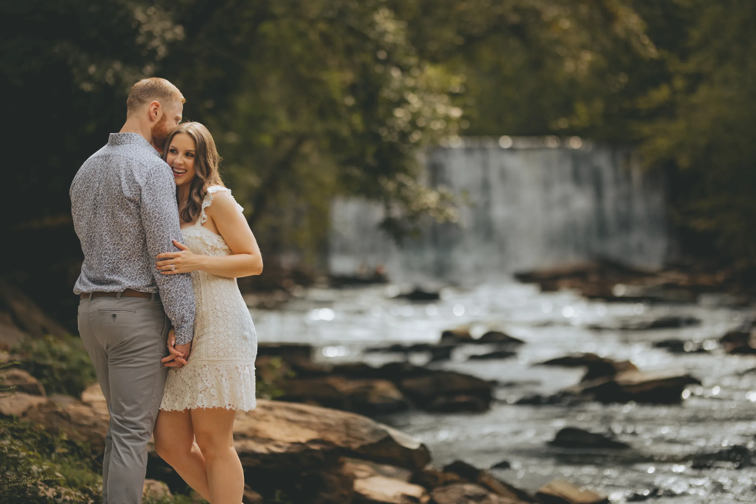 Engagement: smiling by the creek in front of the falls, at their engagement session at Roswell Mill in Roswell, GA