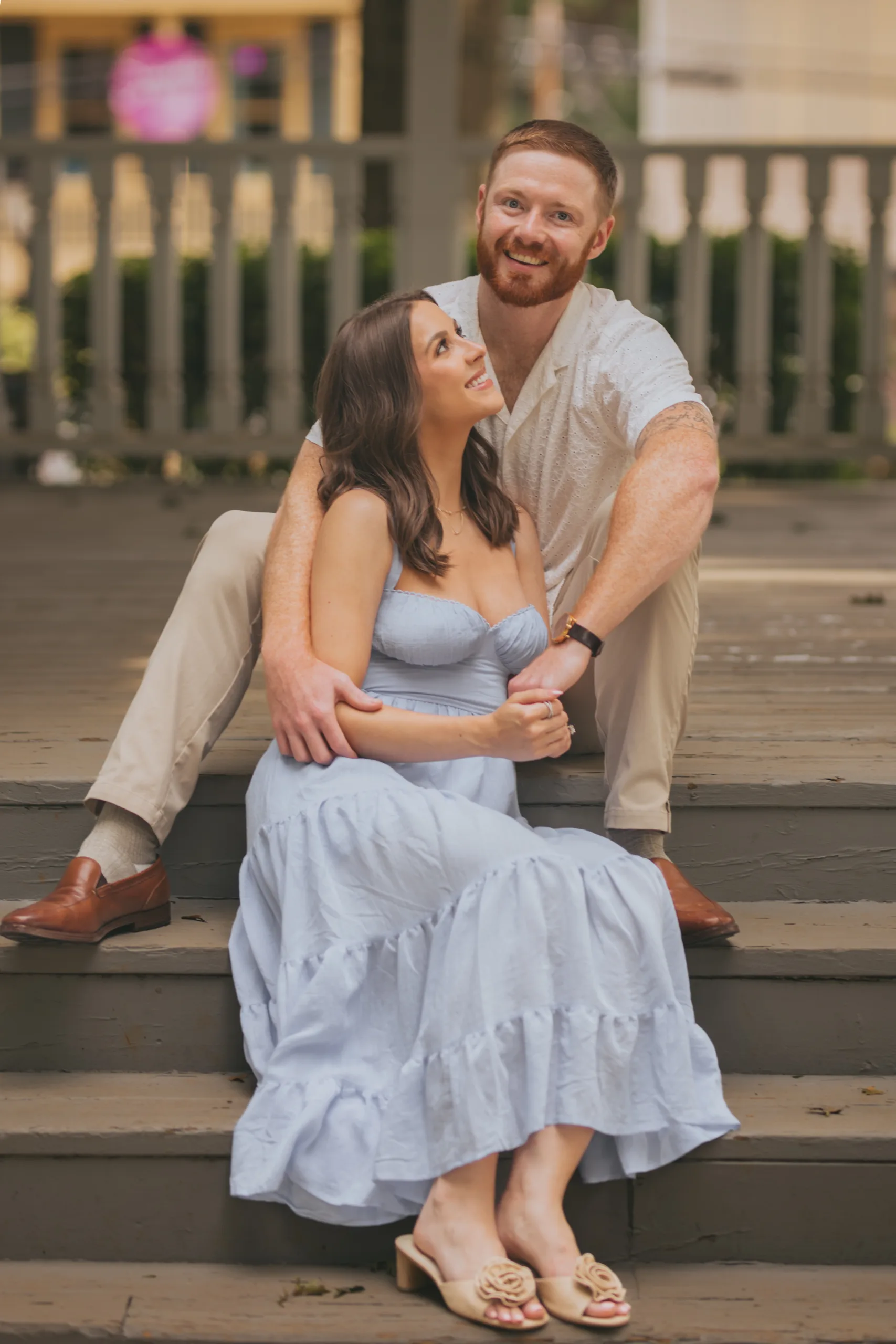 Engagement: sitting together on the steps, her in a blue sundress, at their engagement session on Roswell Square in Roswell, GA