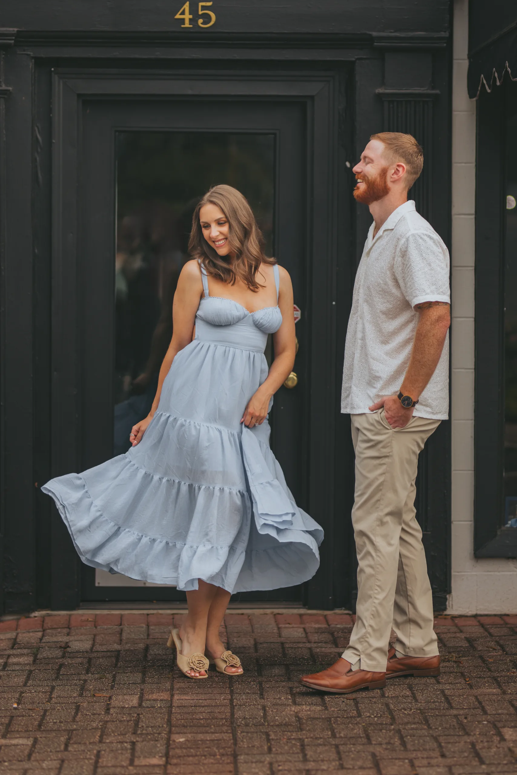 Engagement: her twirling in her sundress by a black door on the sidewalk, at their engagement session on Roswell Square in Roswell, GA