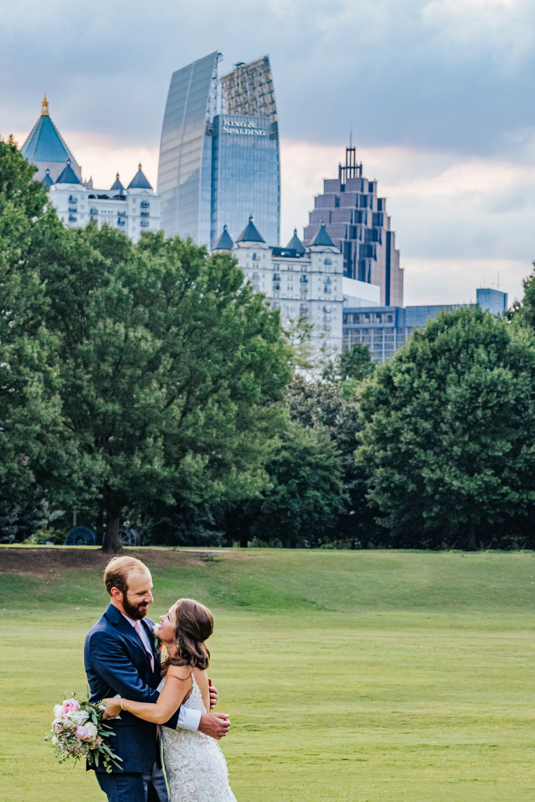 Wedding Formals: an embrace with the Midtown skyline behind them on the meadow in Piedmont Park, at their wedding at The Piedmont Room in Atlanta, GA