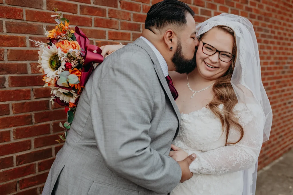 Wedding Formals: a kiss on her cheek against the brick wall, at their wedding at 550 Trackside in Lawrenceville, GA