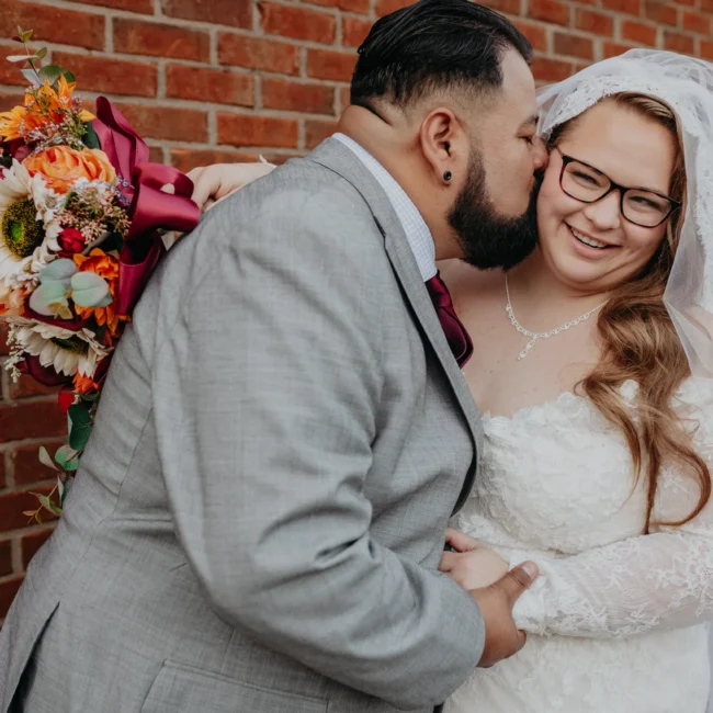 Wedding Formals: a kiss on her cheek against the brick wall, at their wedding at 550 Trackside in Lawrenceville, GA