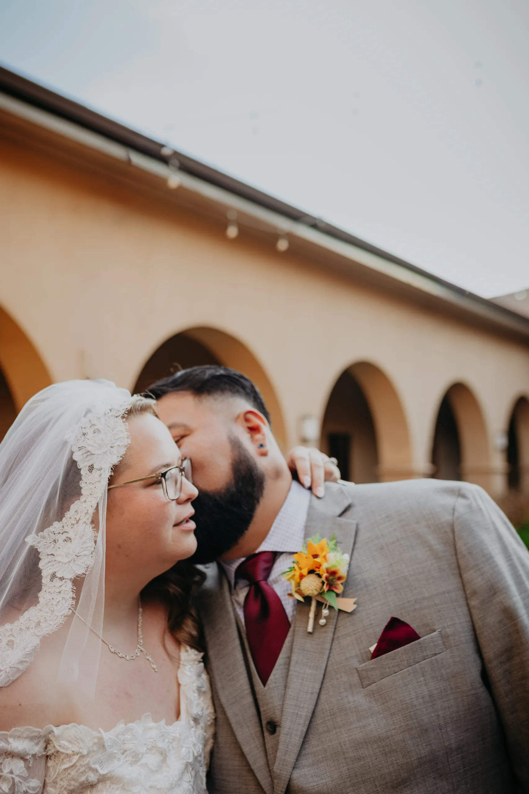 Wedding Formals: a kiss on his cheek under the courtyard arches, at their wedding at St. Lawrence Catholic Church in Lawrenceville, GA