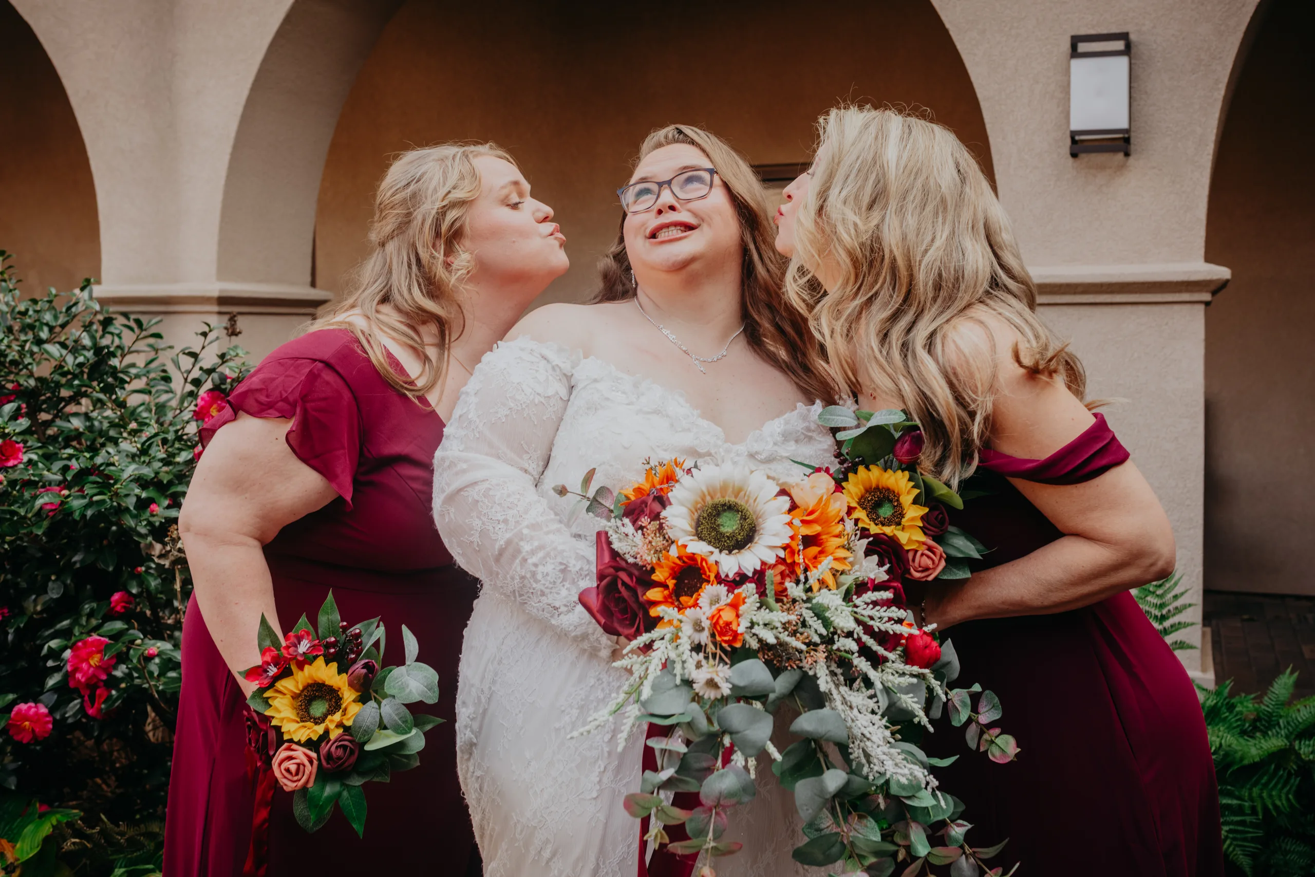 Wedding Party: two bridesmaids kissing her cheeks in the courtyard, at their wedding at St. Lawrence Catholic Church in Lawrenceville, GA