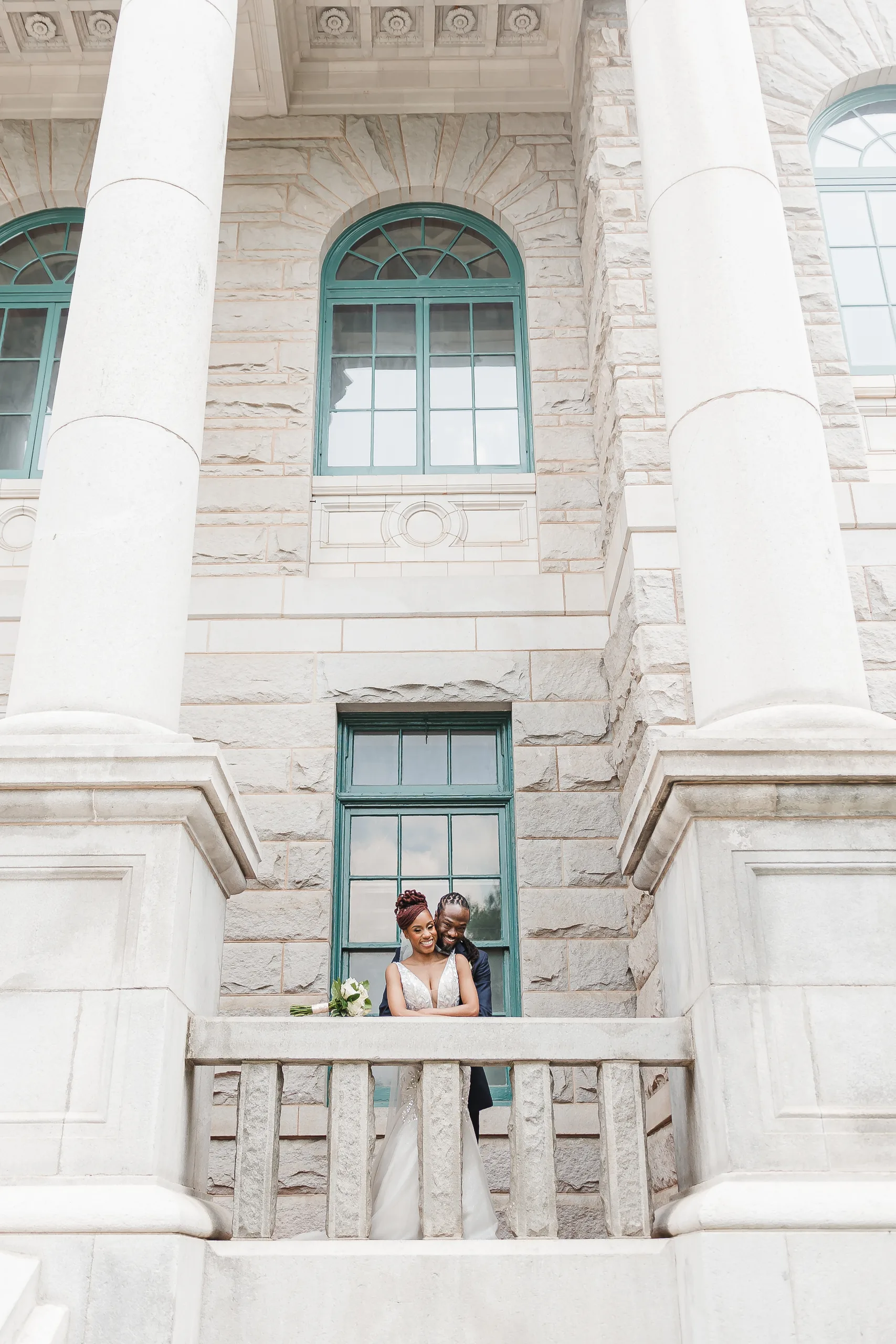 Wedding Formals: the two of them on the balcony between the tall stone columns, at their wedding at the Historic DeKalb Courthouse in Decatur, GA