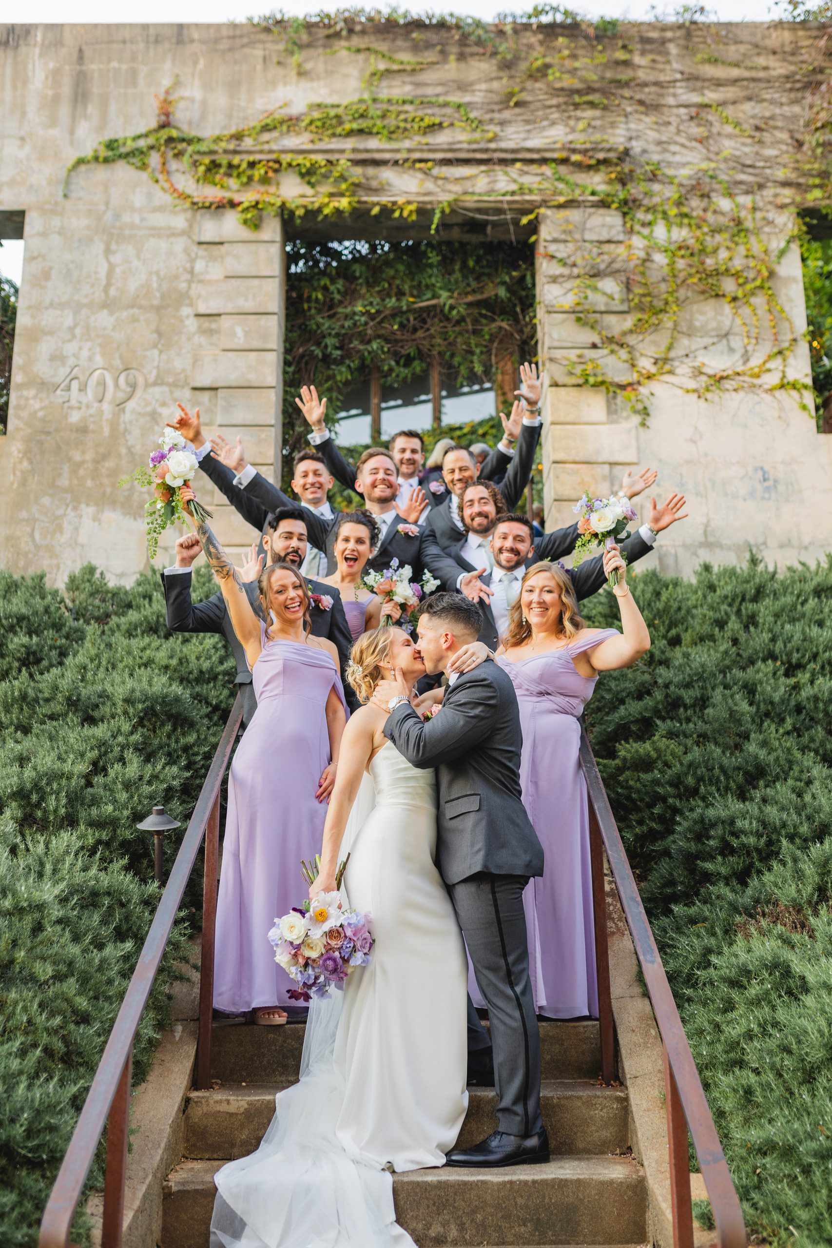 Wedding Party: the two of them kissing on the steps as the party cheers behind them, at their wedding at Summerour Studio in Atlanta, GA