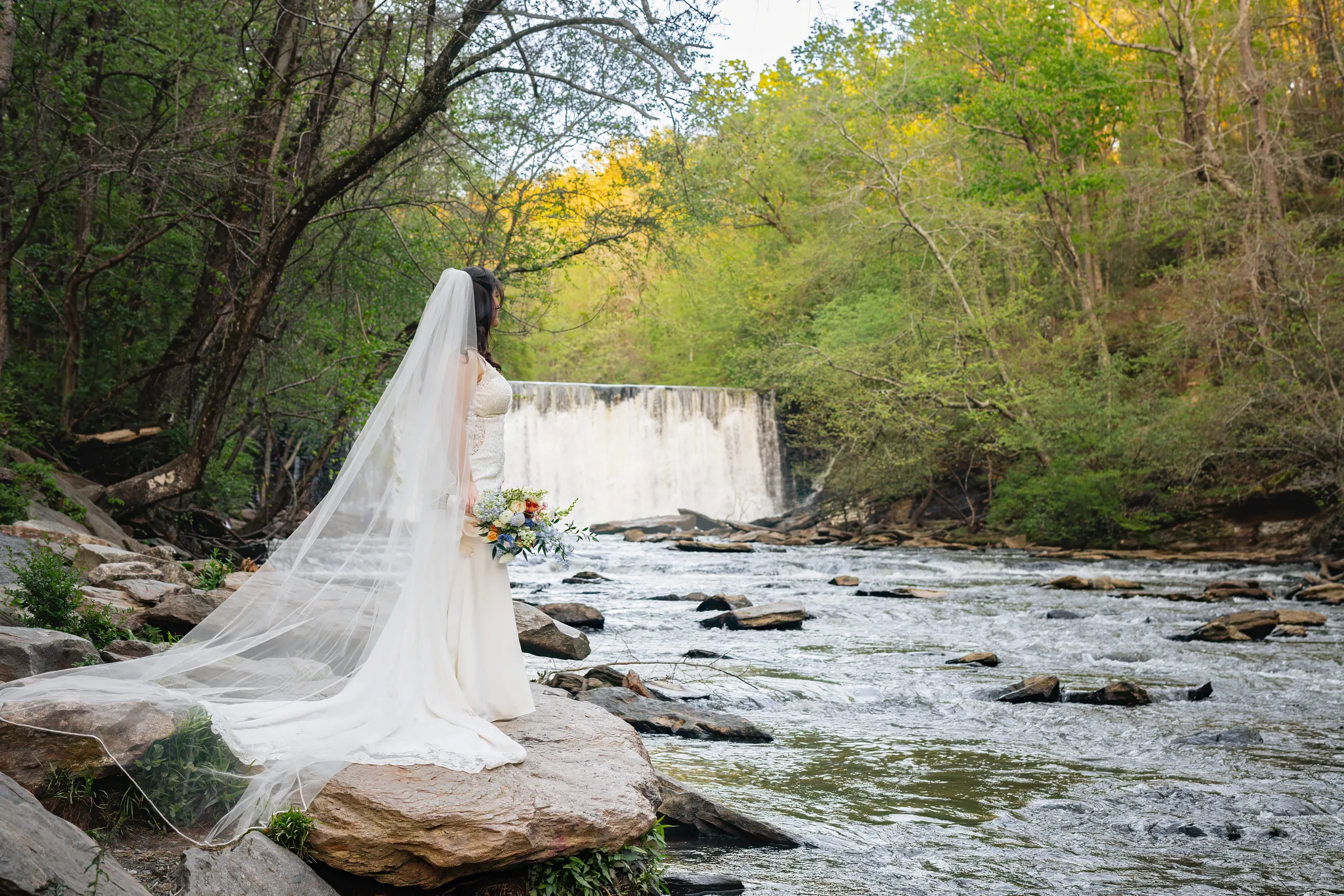Bridal Portraits: the bride with her veil spread across the rocks by the falls at their wedding at Ivy Hall at Roswell Mill in Roswell, GA
