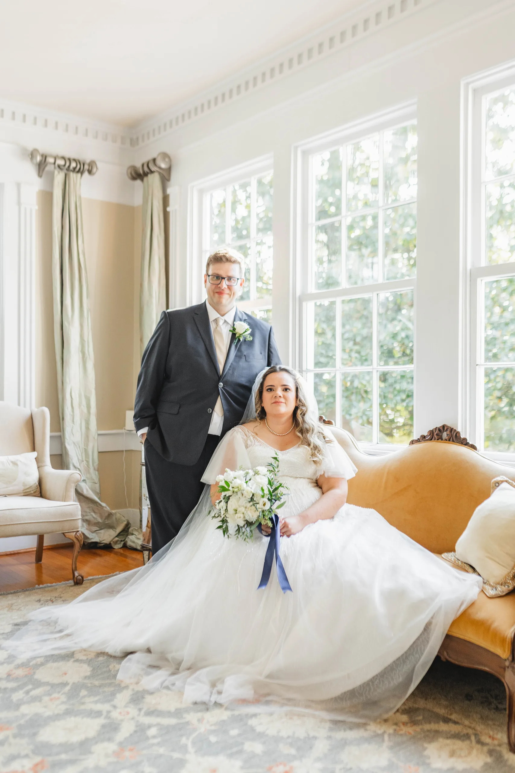 Wedding Formals: the bride on the gold settee, the groom standing beside her by the windows, at their wedding at Naylor Hall in Roswell, GA