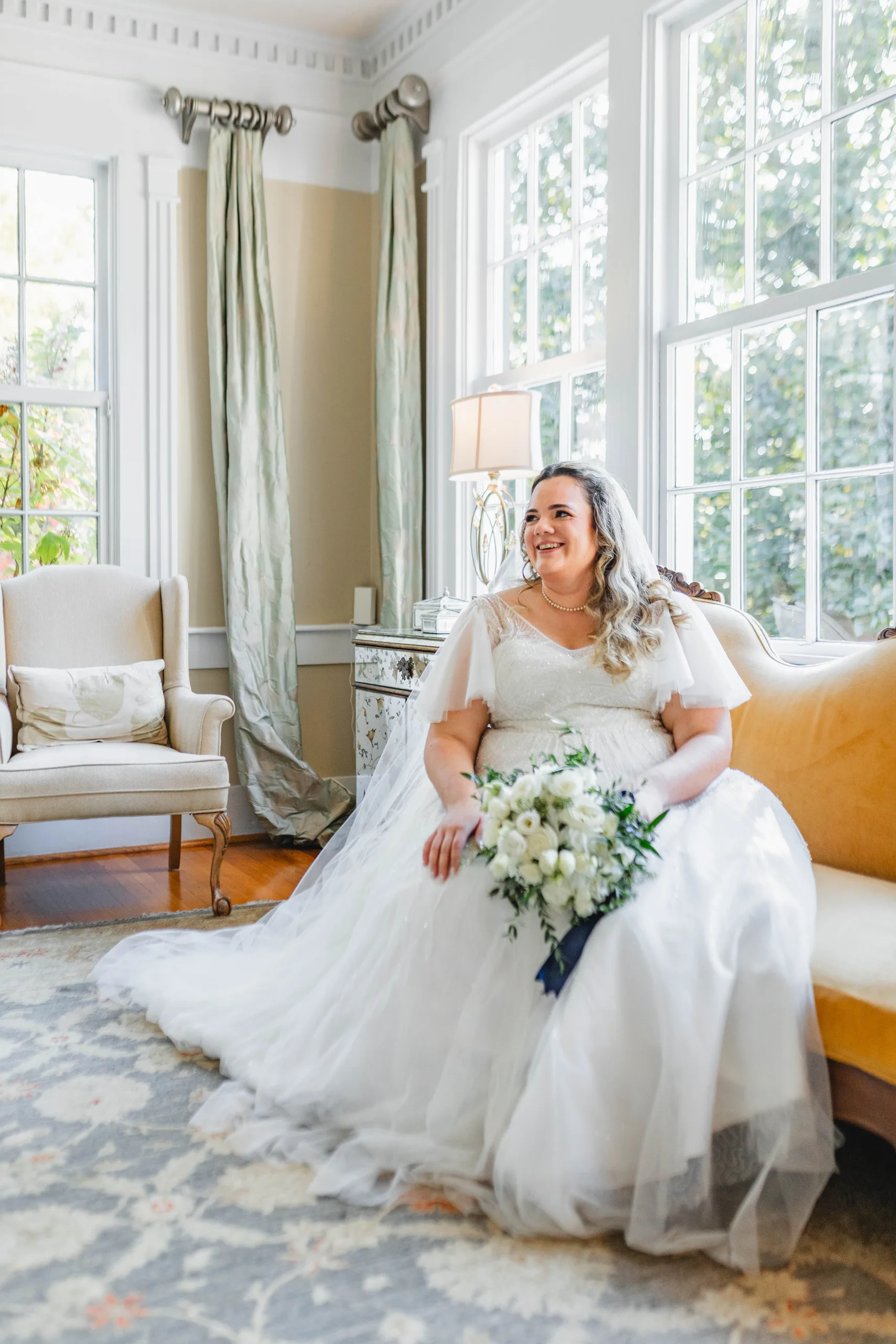 Bridal Portraits: the bride laughing on the gold settee by the windows, at their wedding at Naylor Hall in Roswell, GA
