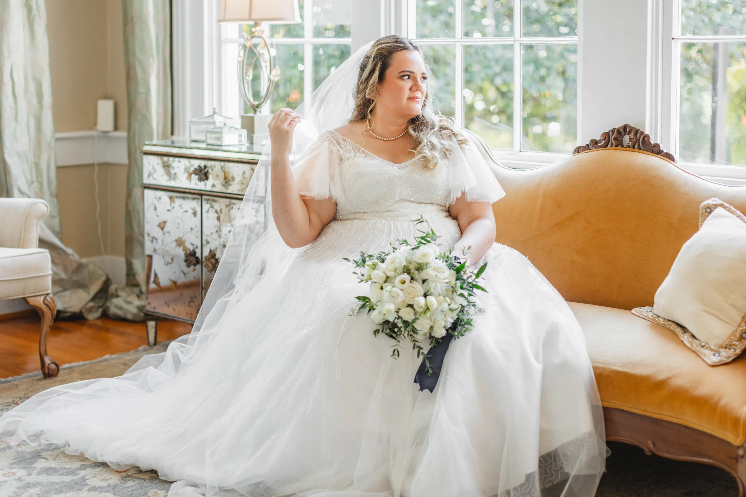 Bridal Portraits: the bride on the gold settee holding her veil, looking out the window, at their wedding at Naylor Hall in Roswell, GA