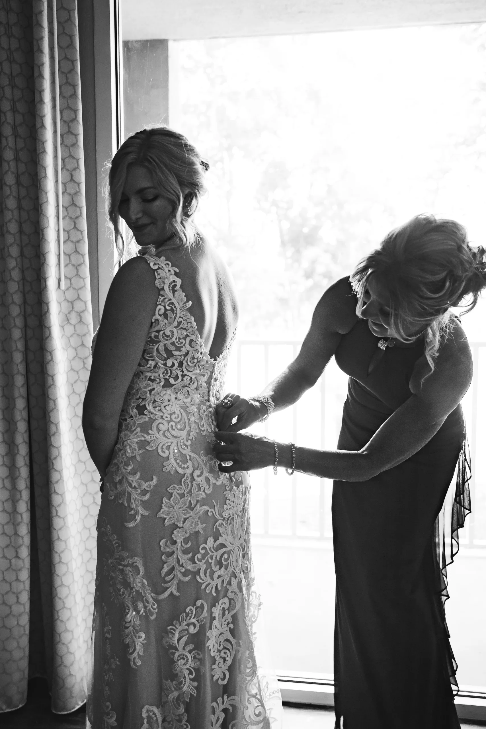 Getting Ready: her mom buttoning up the back of her lace gown, in black and white, before their wedding at Atlanta Evergreen Lakeside Resort in Stone Mountain, GA