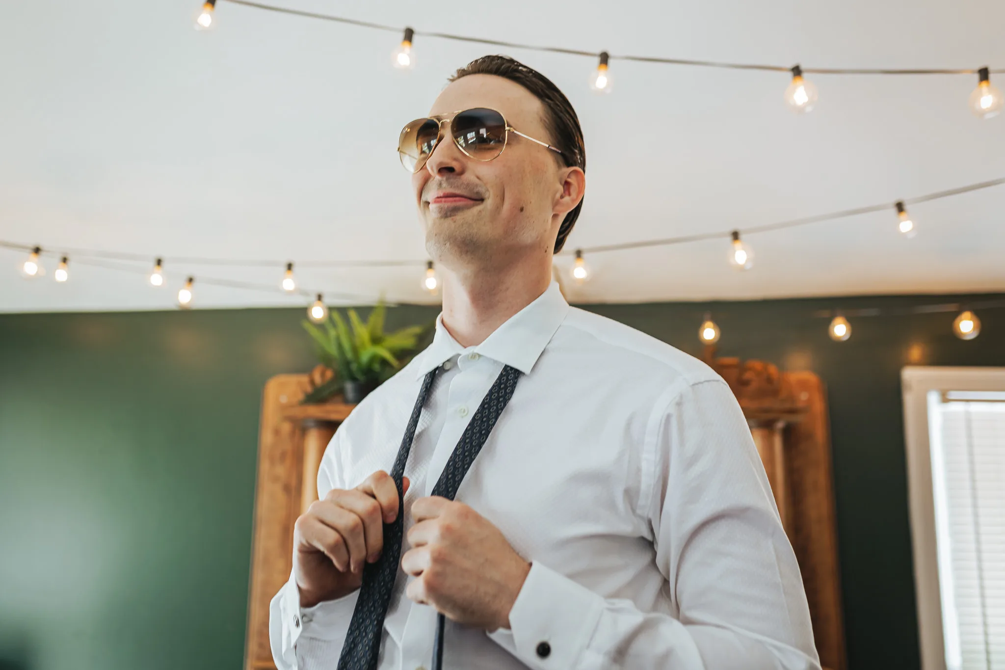 Getting Ready: the groom in sunglasses tying his tie under string lights, before their wedding at The Wildflower 301 in Greensboro, GA