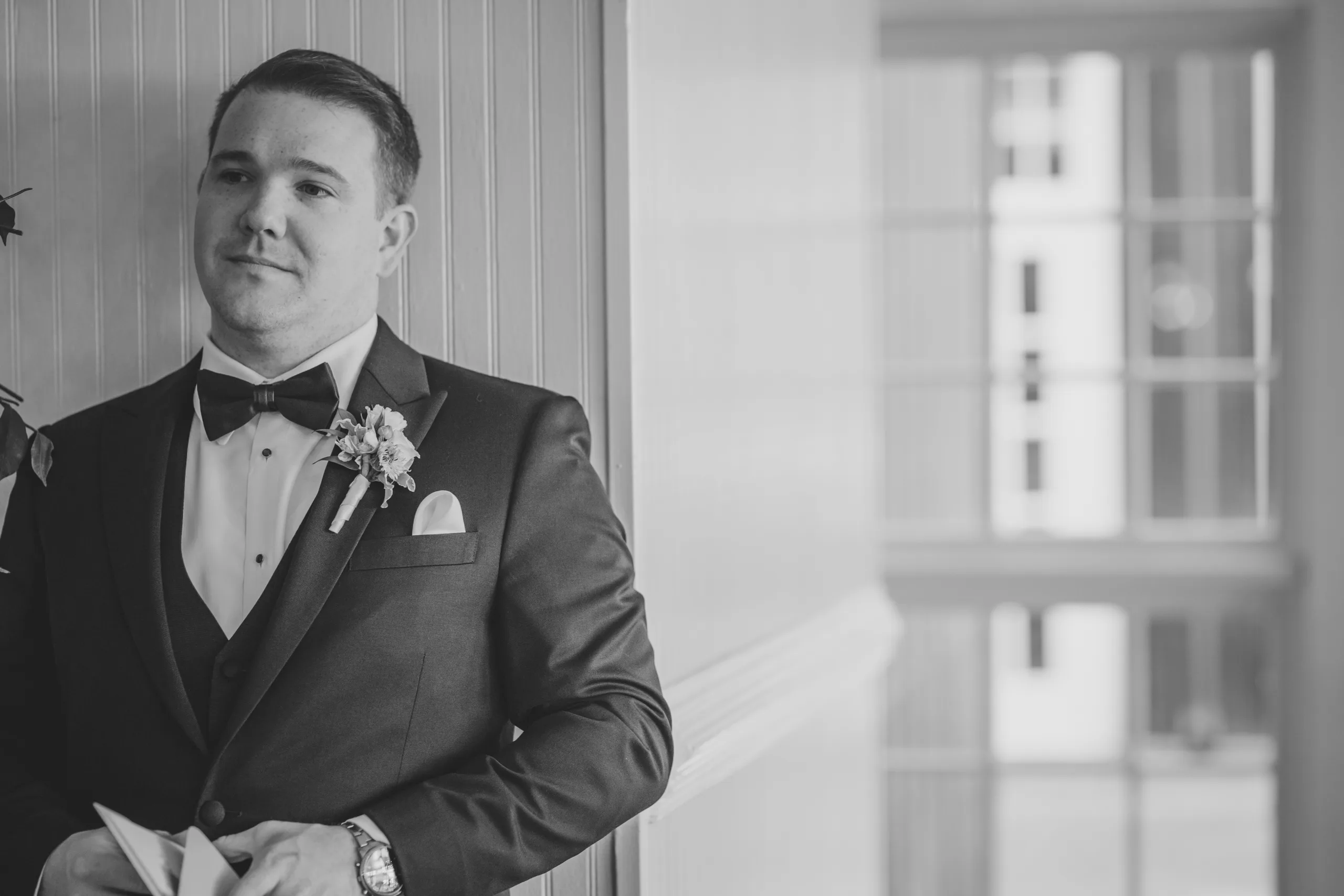 Getting Ready: the groom smiling as he reads, in black and white before their wedding at Ivy Hall at Roswell Mill in Roswell, GA
