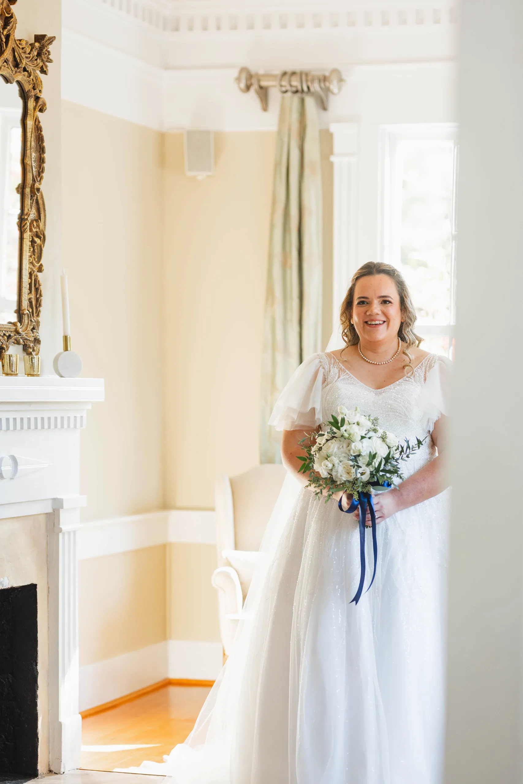 Getting Ready: the bride in her gown with her bouquet, by the fireplace and gilded mirror, before their wedding at Naylor Hall in Roswell, GA